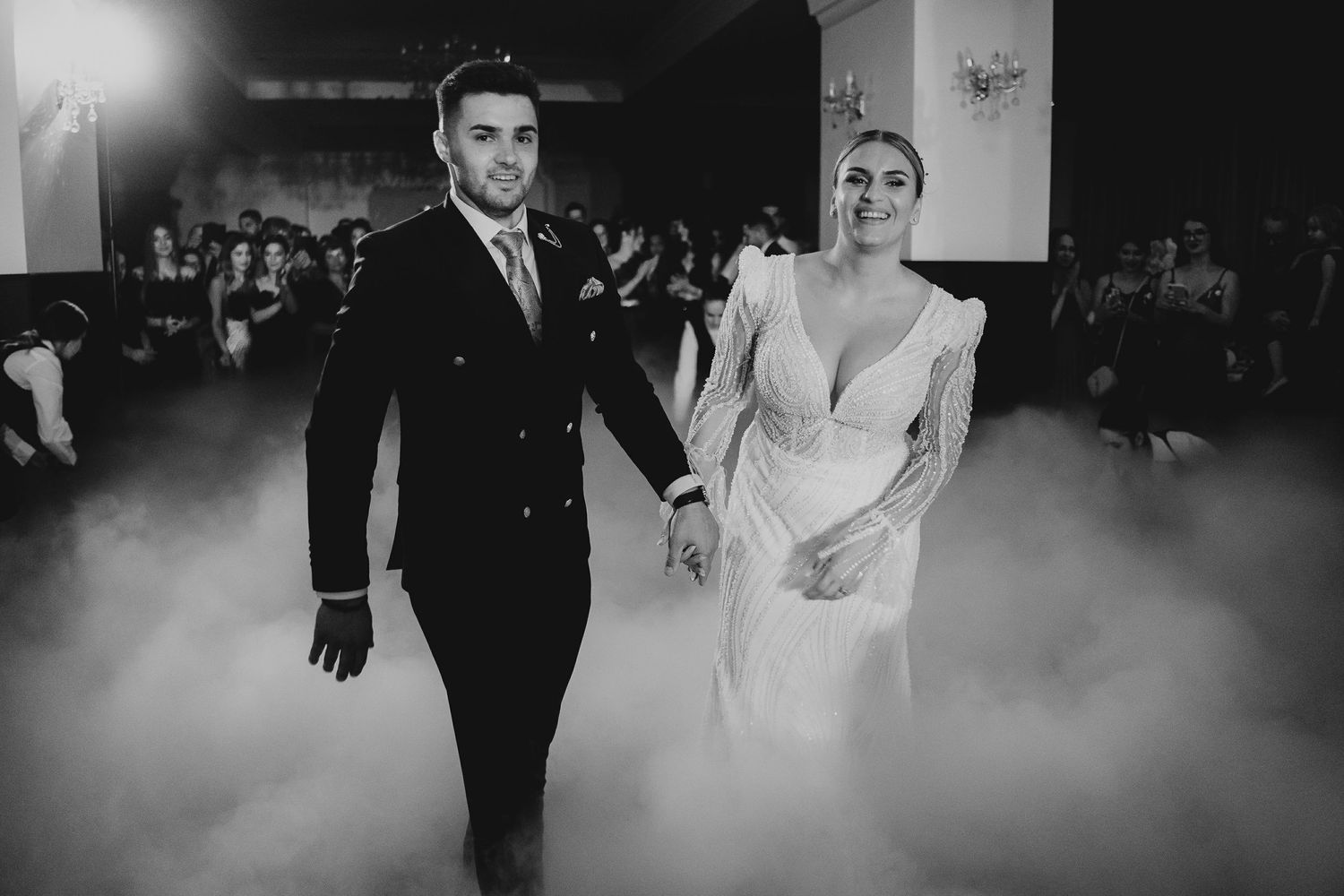Bride and groom walk hand in hand through fog during wedding reception entrance in black and white.