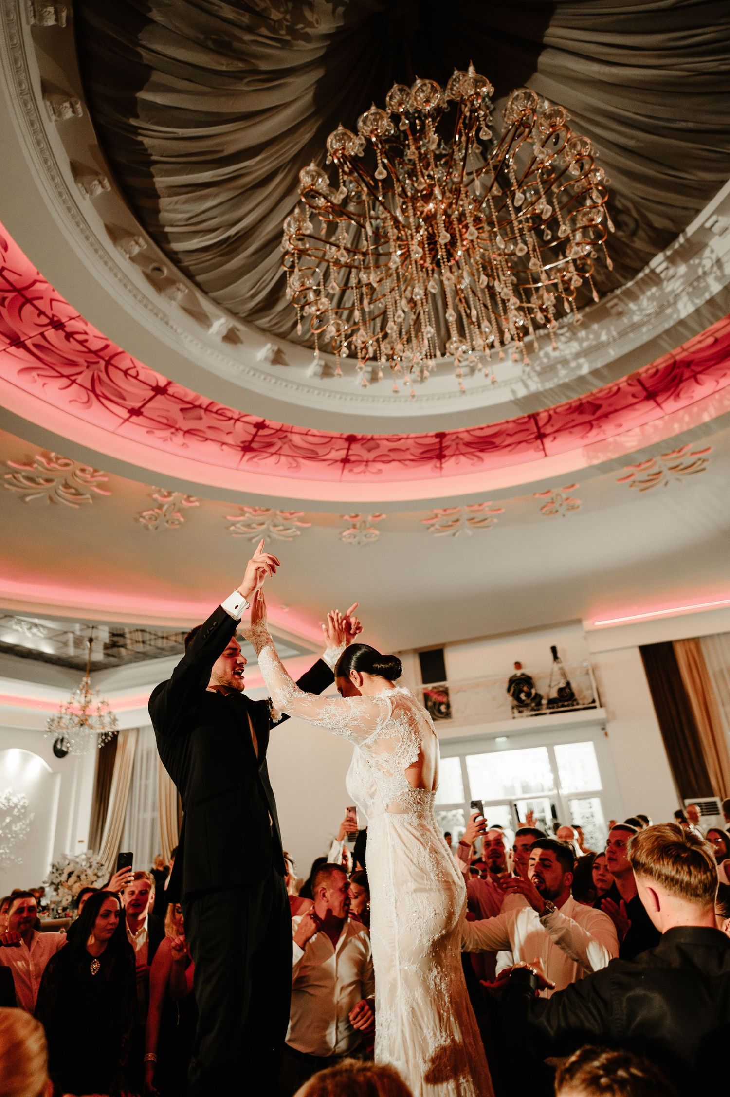 Bride and groom dancing under a crystal chandelier at an elegant wedding reception with pink lighting.