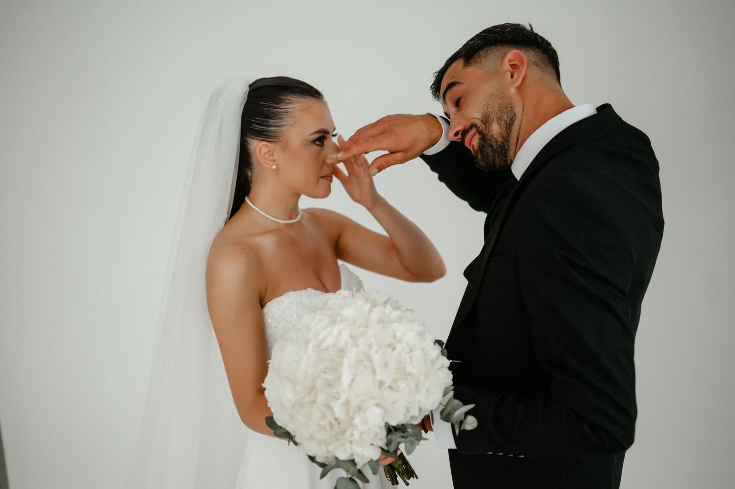 Groom in black suit gently kisses bride's hand as she smiles holding a white hydrangea bouquet.