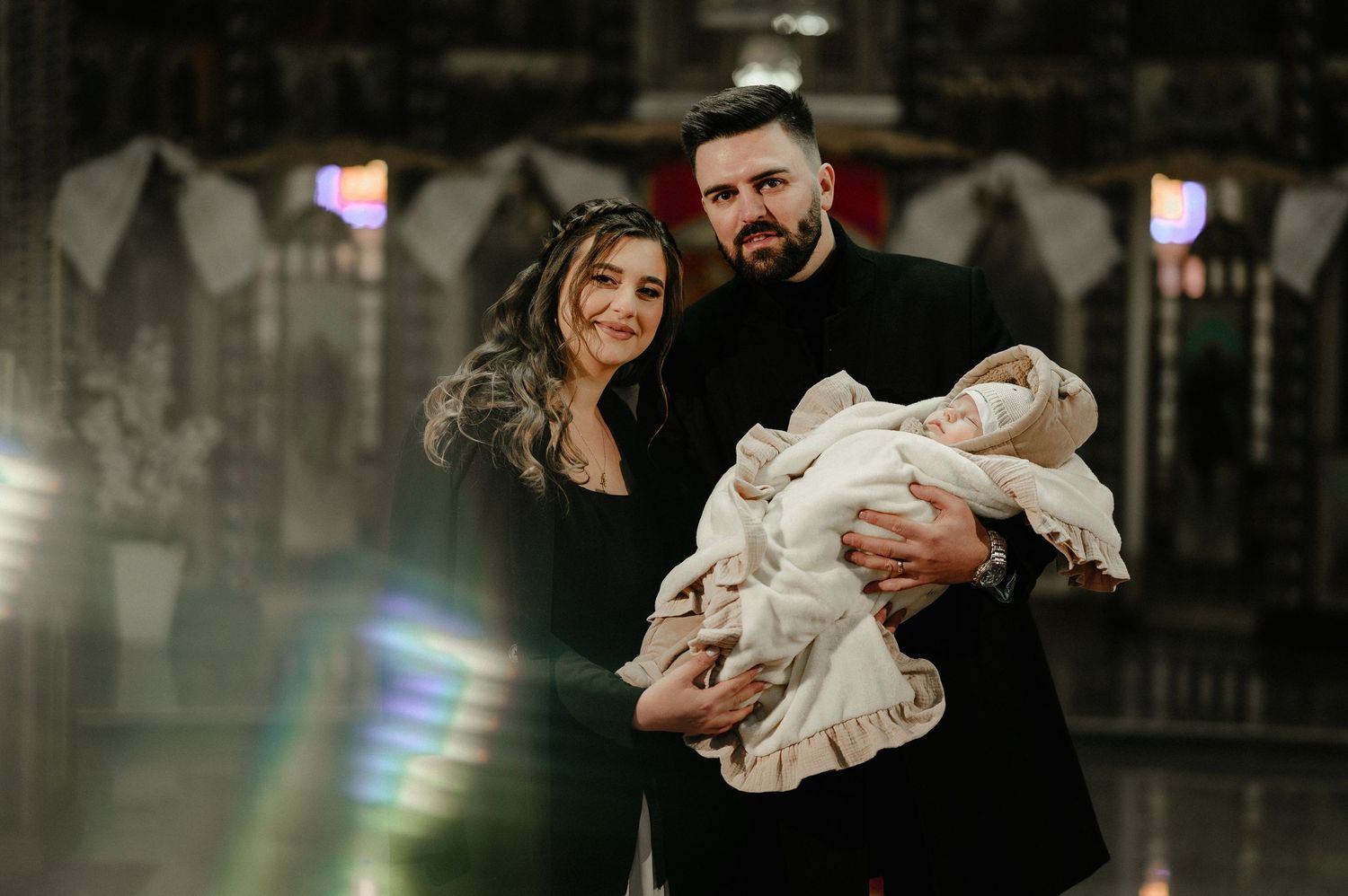 Young couple in black attire holding newborn baby wrapped in white baptism cloth.