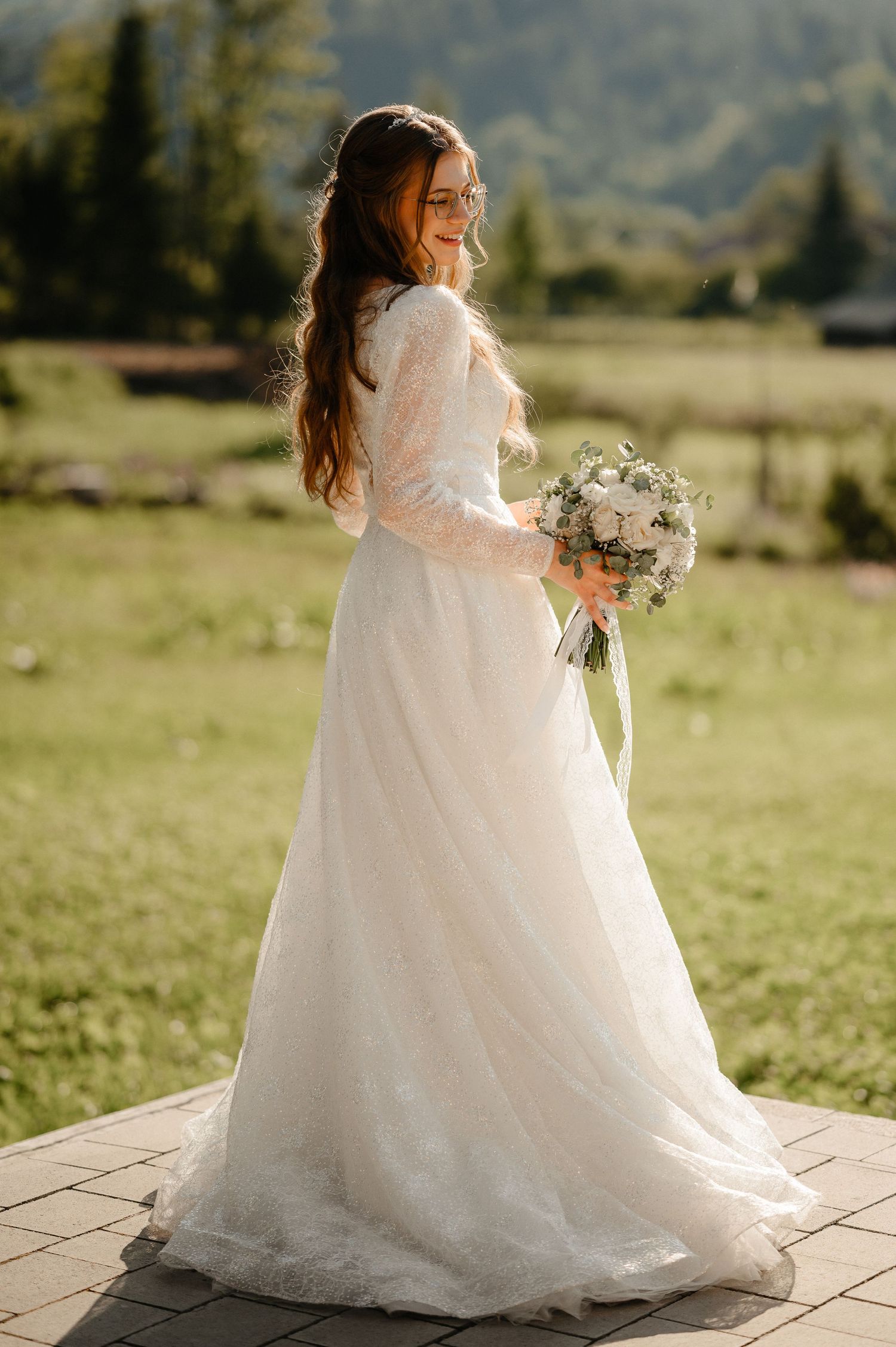 Bride in elegant long-sleeve white lace wedding gown holding bouquet outdoors with mountain backdrop.