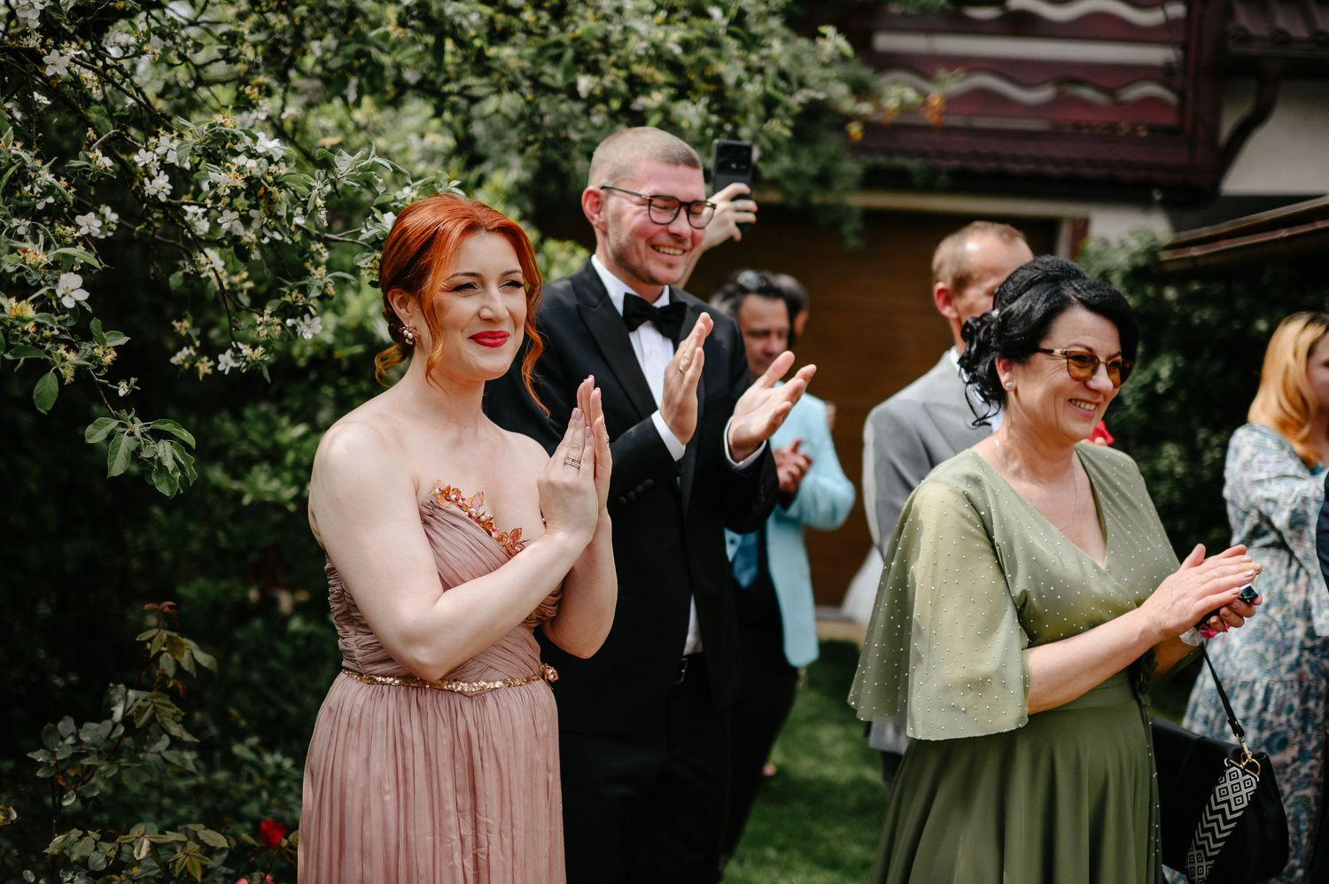 Wedding guests in elegant attire clap and celebrate outdoors near blooming white trees at reception.
