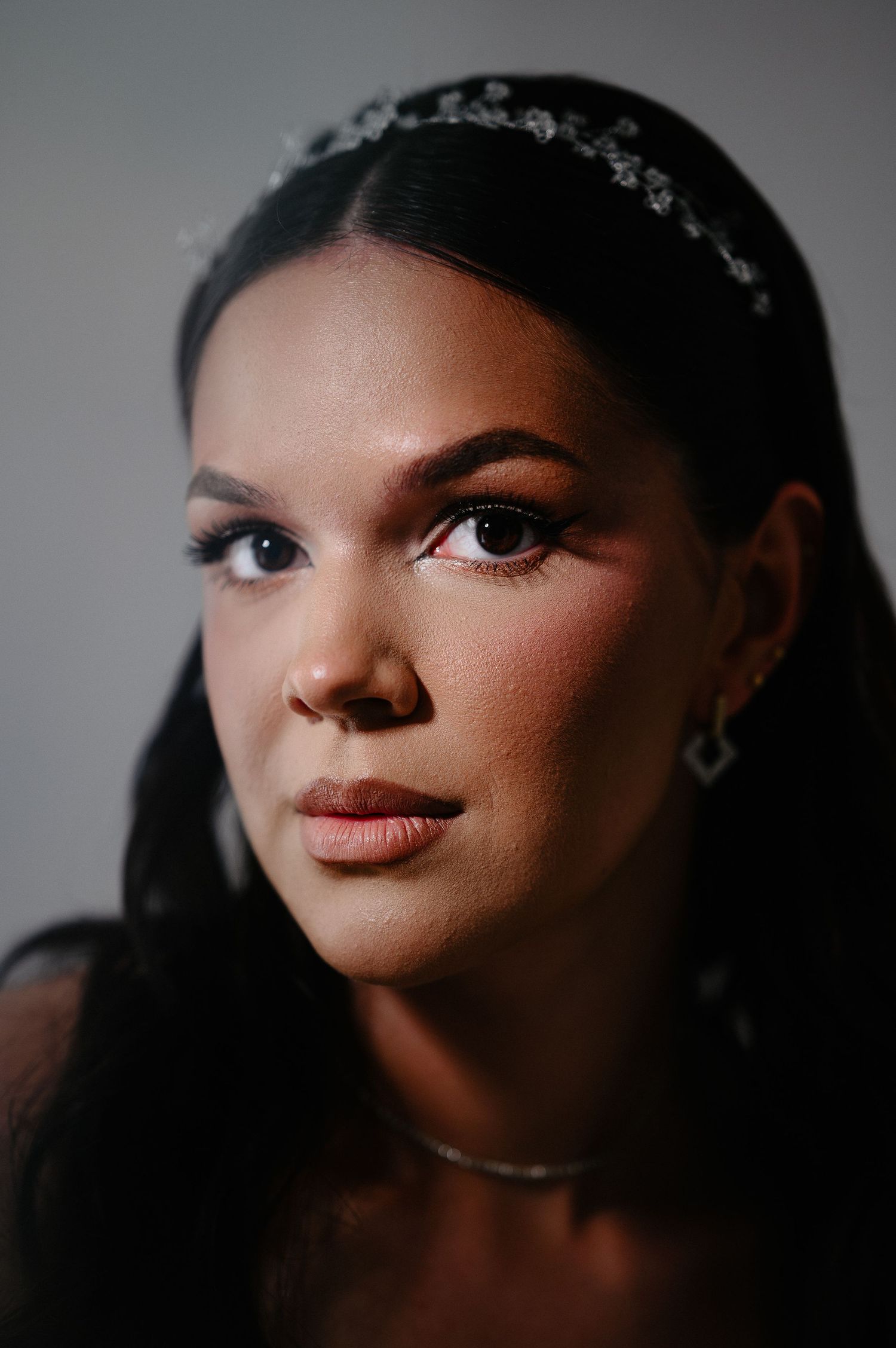 Close-up portrait of dark-haired bride wearing crystal headband, diamond earrings, and delicate necklace.