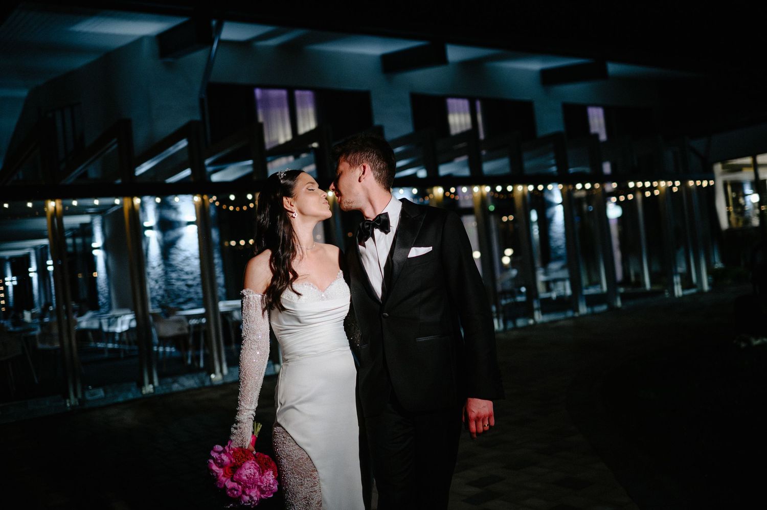 Bride and groom sharing a romantic kiss outdoors at night with string lights at wedding venue.