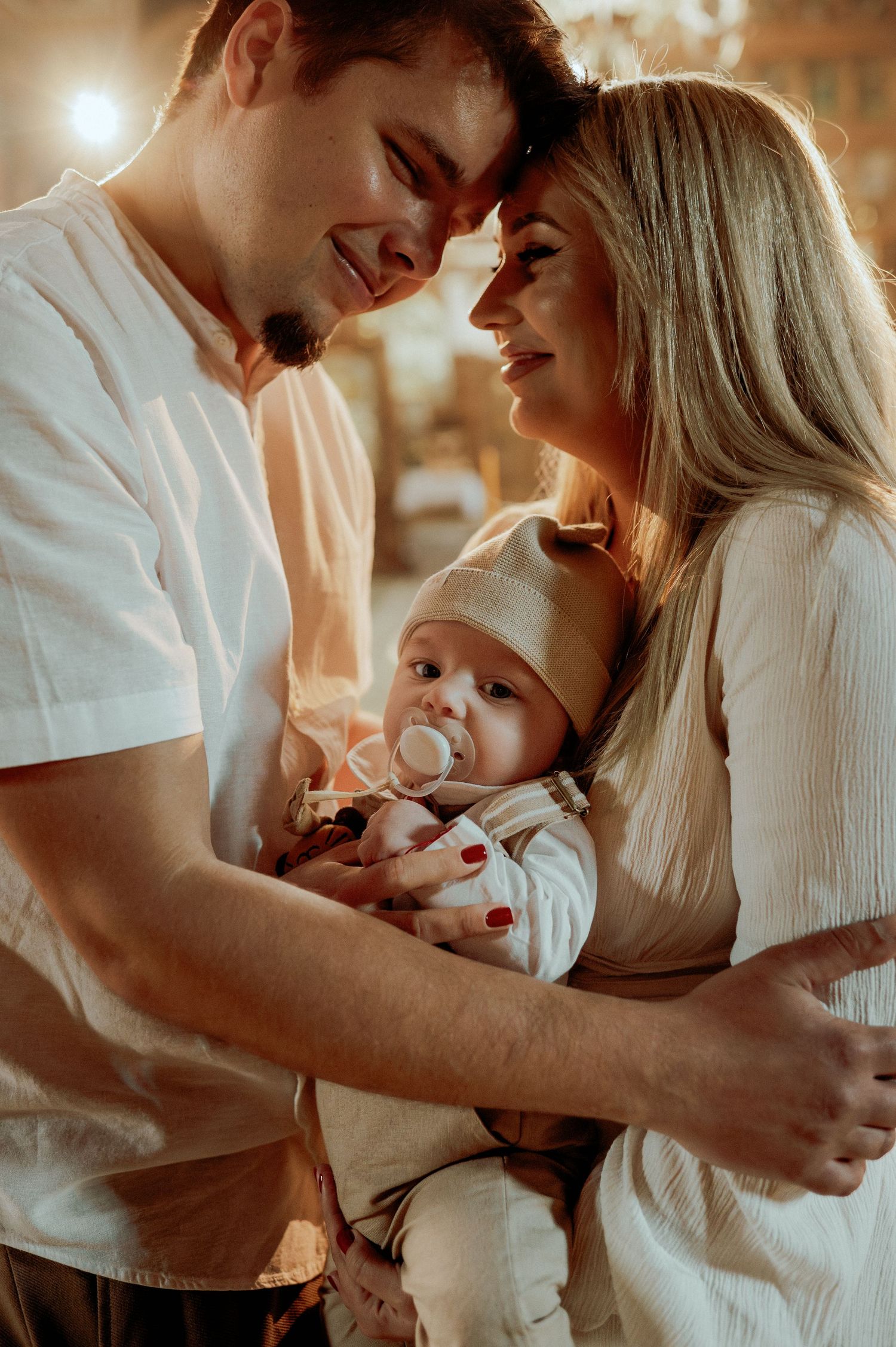 Happy couple touching foreheads while holding baby with pacifier in warm light.
