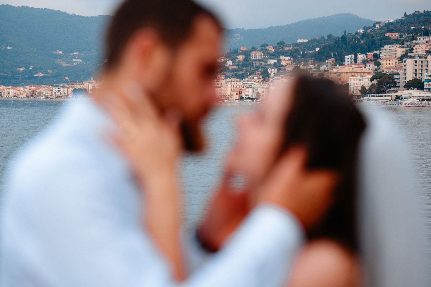 Blurred romantic couple kissing with scenic coastal town visible in background.