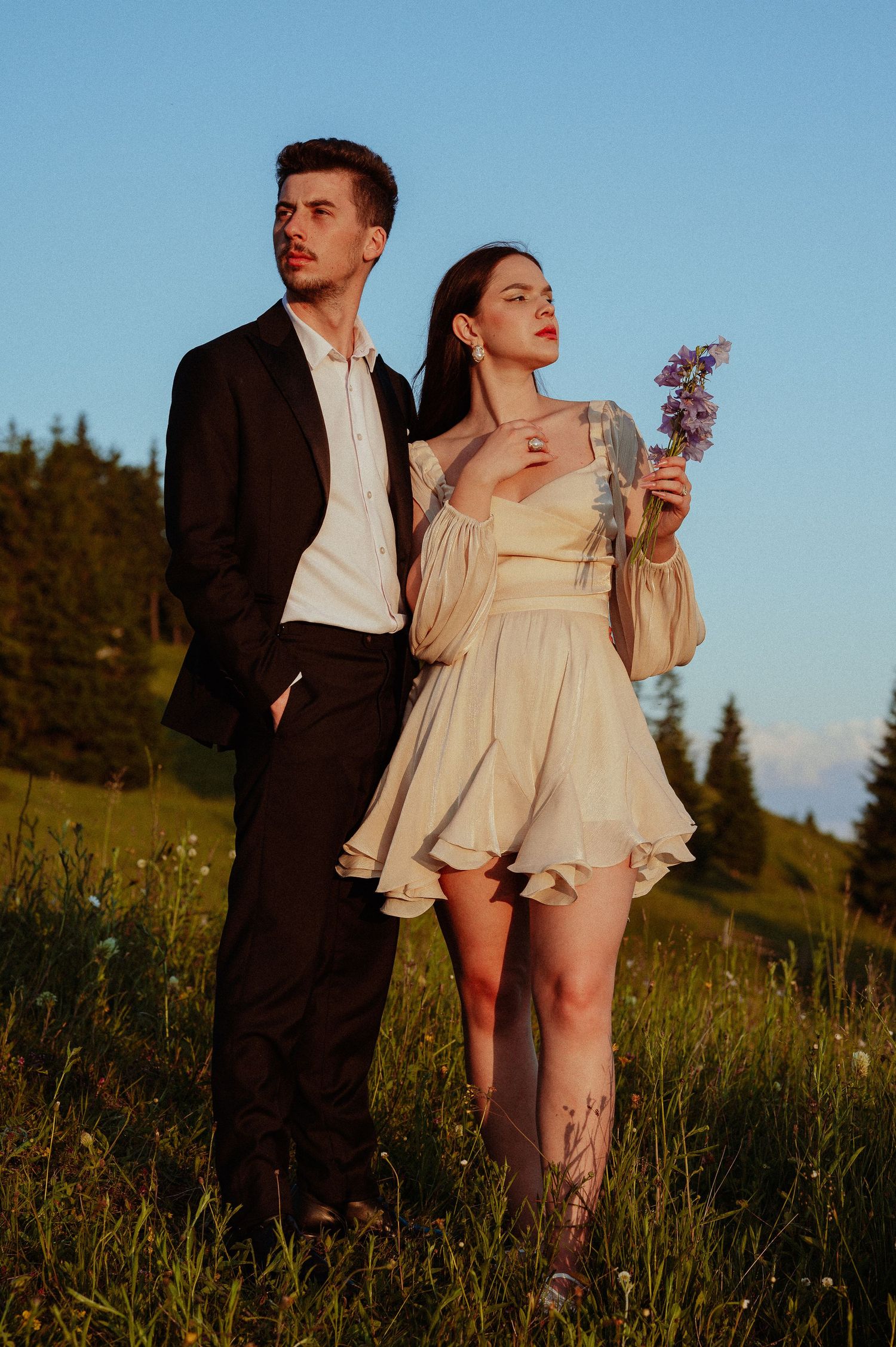 Couple standing in a green meadow at sunset, man in black suit, woman in cream mini dress holding purple wildflowers.