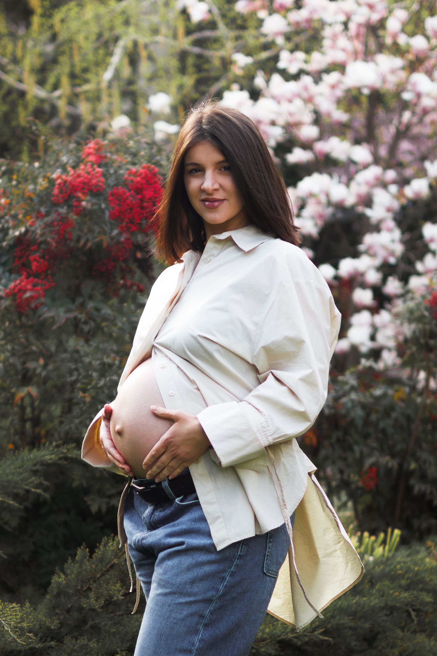 Pregnant woman in white shirt and jeans holding baby bump outdoors near blooming flowers.