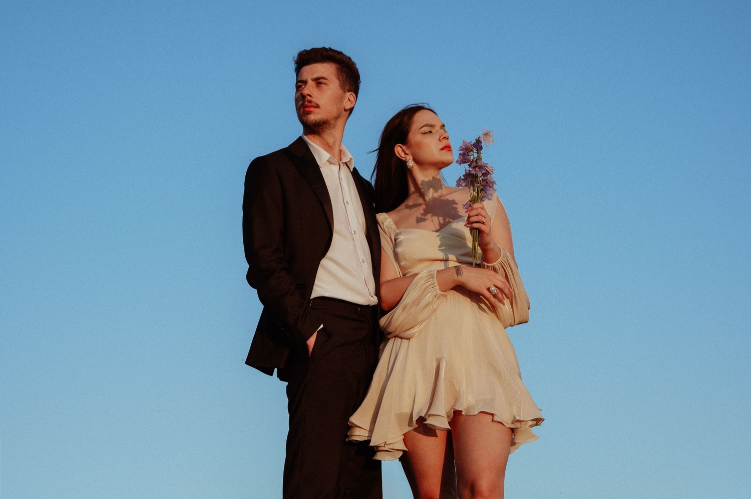 A stylish couple poses against a blue sky, man in black suit, woman in beige dress holding flowers.