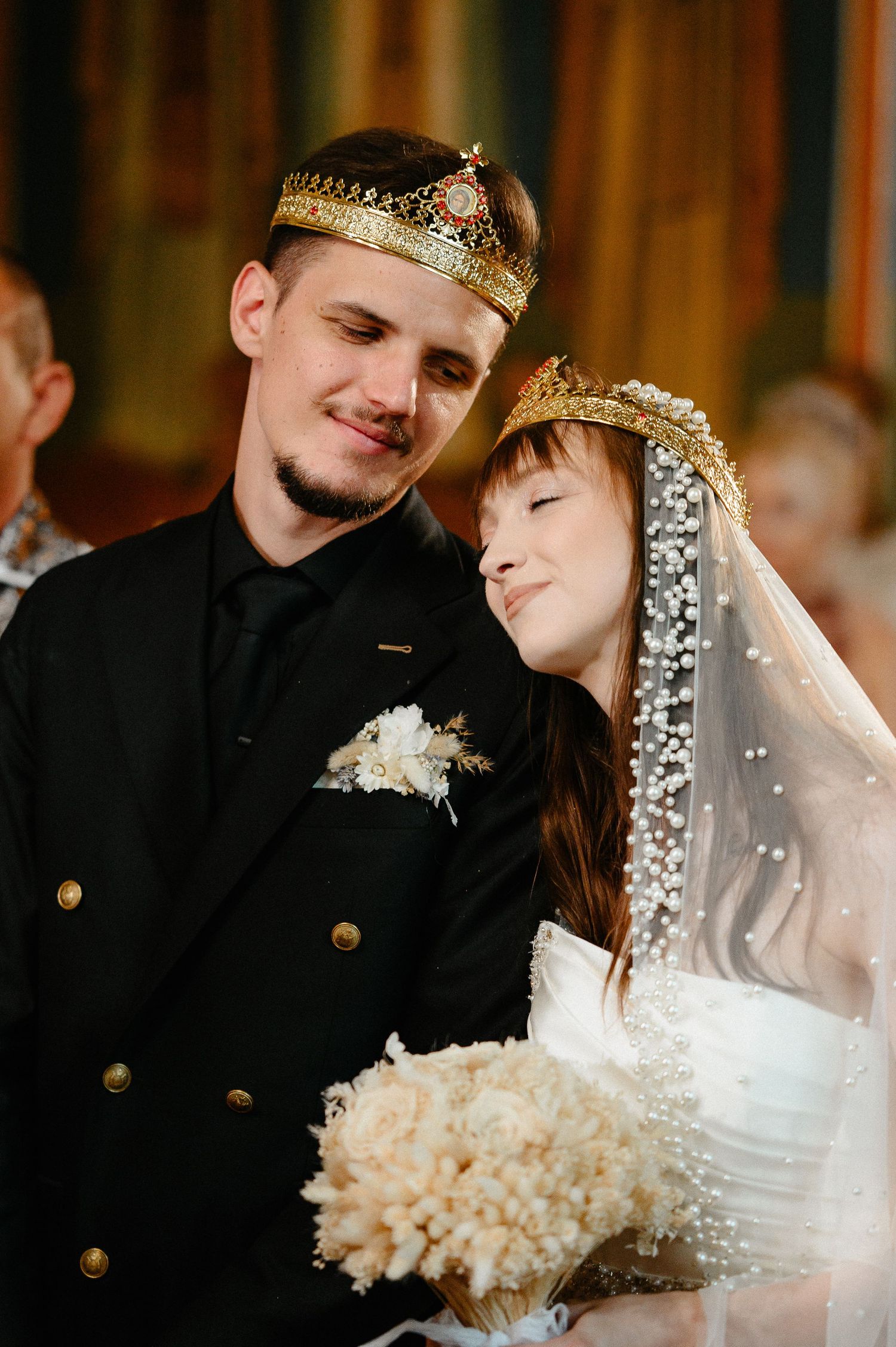 Bride and groom wearing traditional Orthodox gold crowns share tender moment during church ceremony.