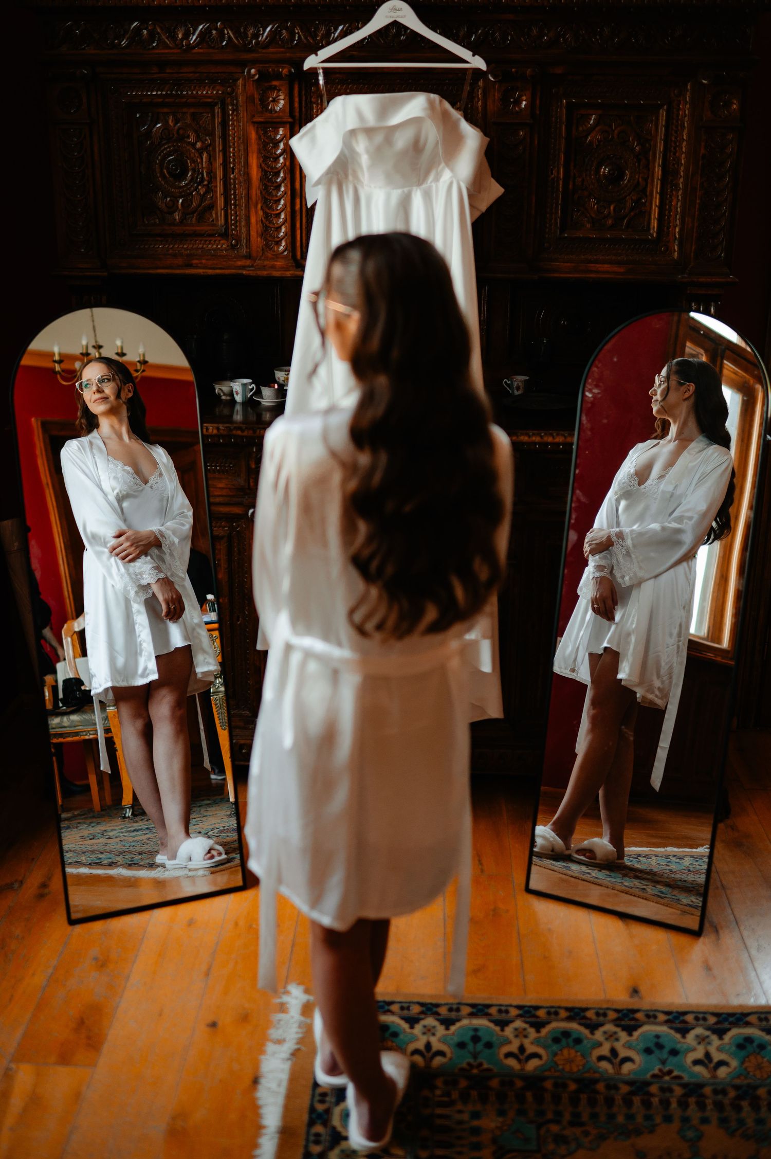 Bride in white robe gazing at her wedding dress hanging between two mirrors in elegant room.
