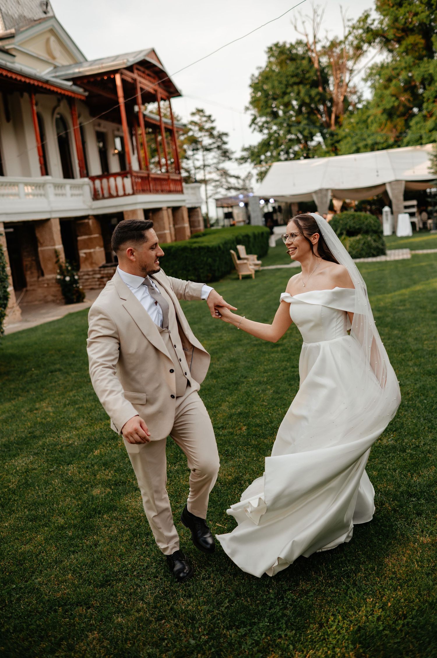 Joyful bride and groom dancing on green lawn with elegant venue building in background.