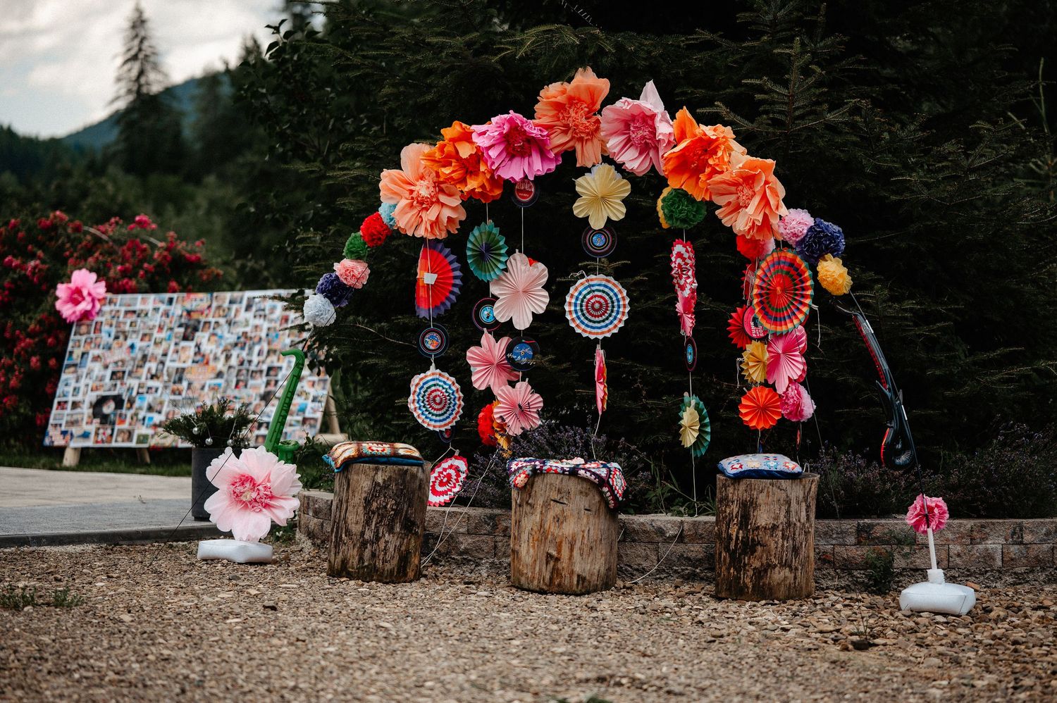 Colorful floral arch with paper flowers and decorations set outdoors among trees for a festive event.