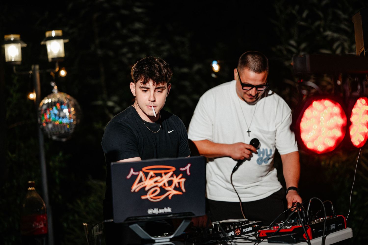 Two DJs mixing music at outdoor night party behind illuminated DJ booth with red stage lights.