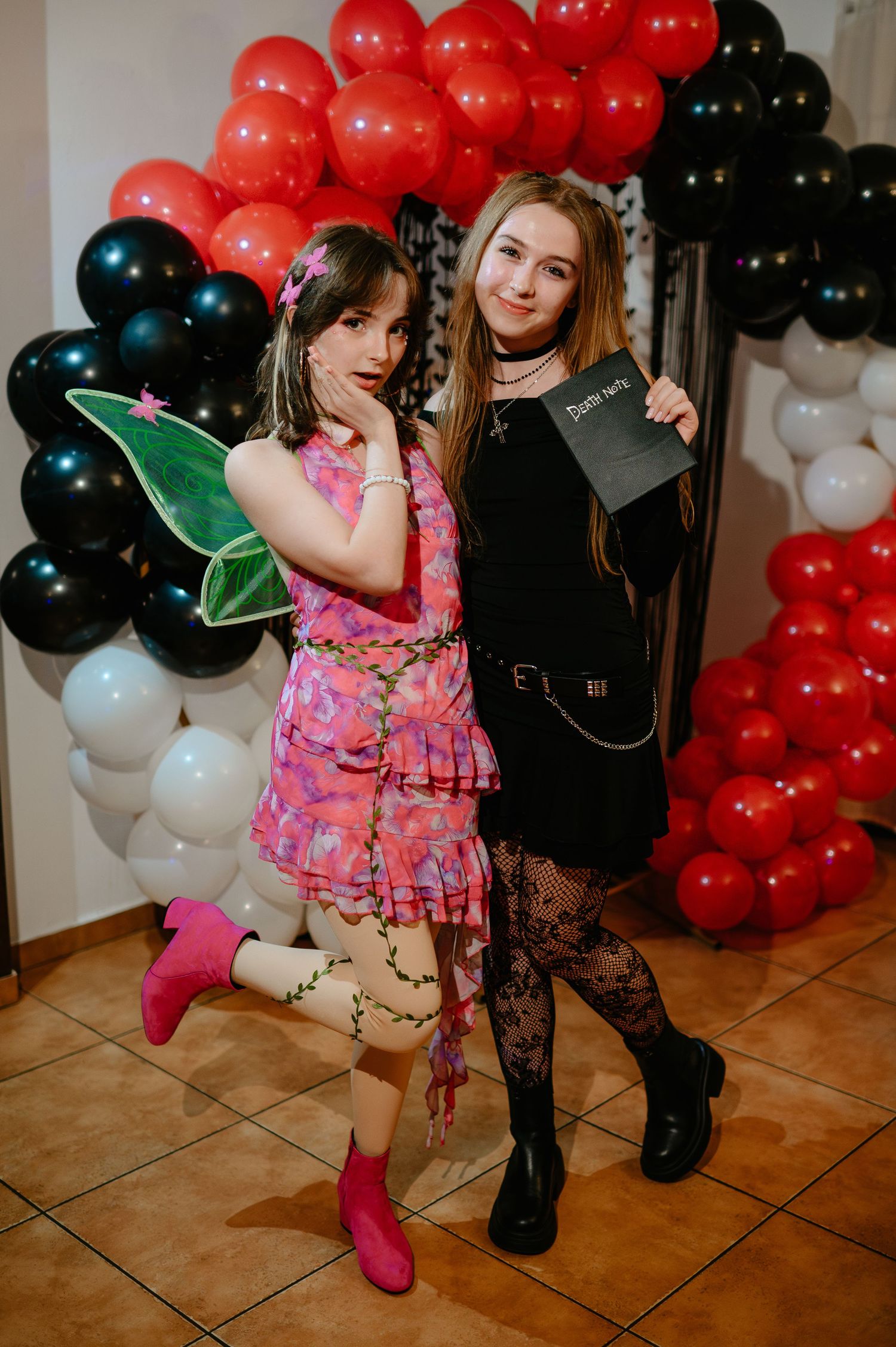 Two women in Halloween costumes pose before a red, black, and white balloon arch at a festive party.