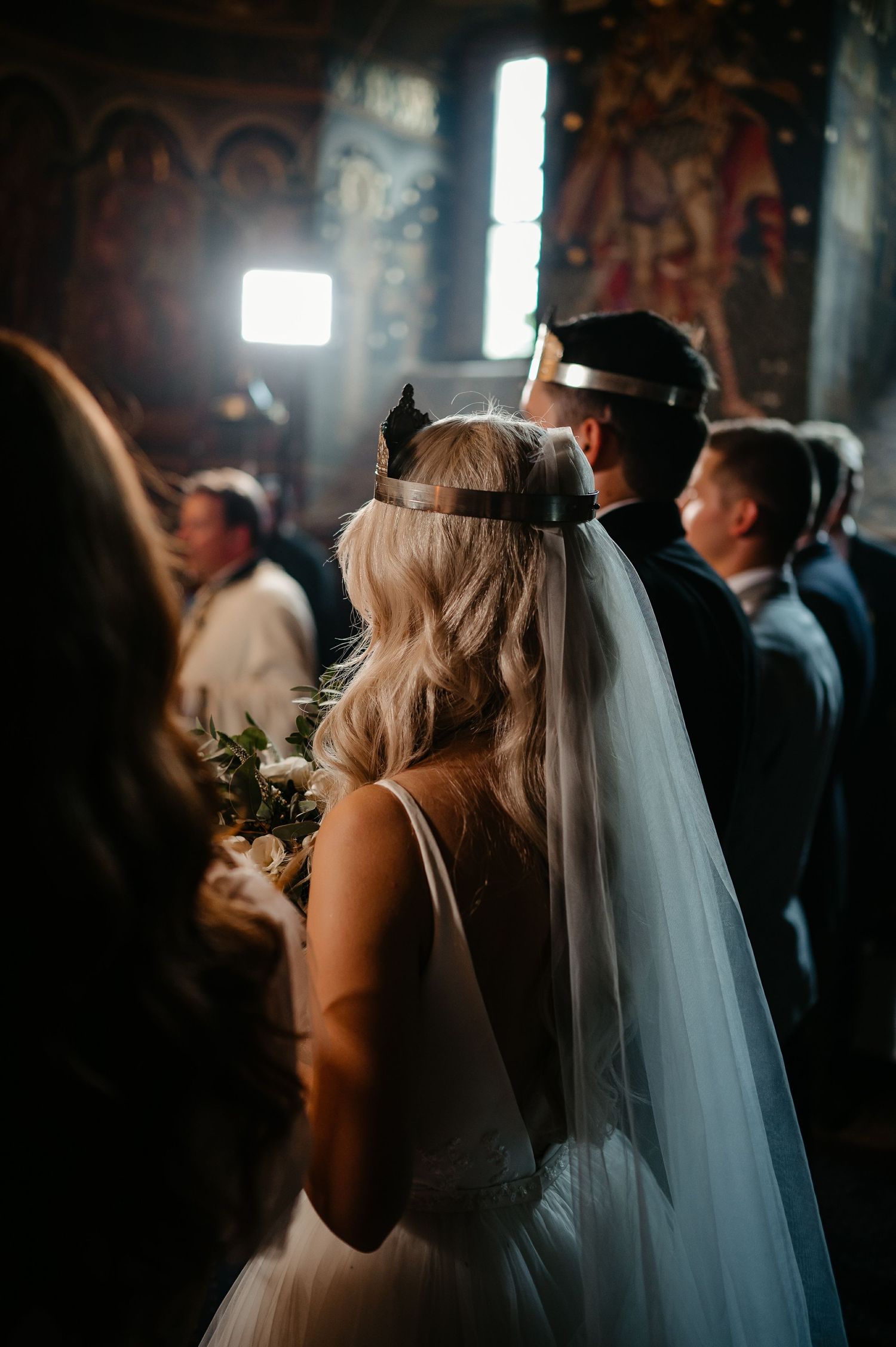 Bride wearing ceremonial crown and veil holding bouquet from behind during Orthodox church wedding ceremony.