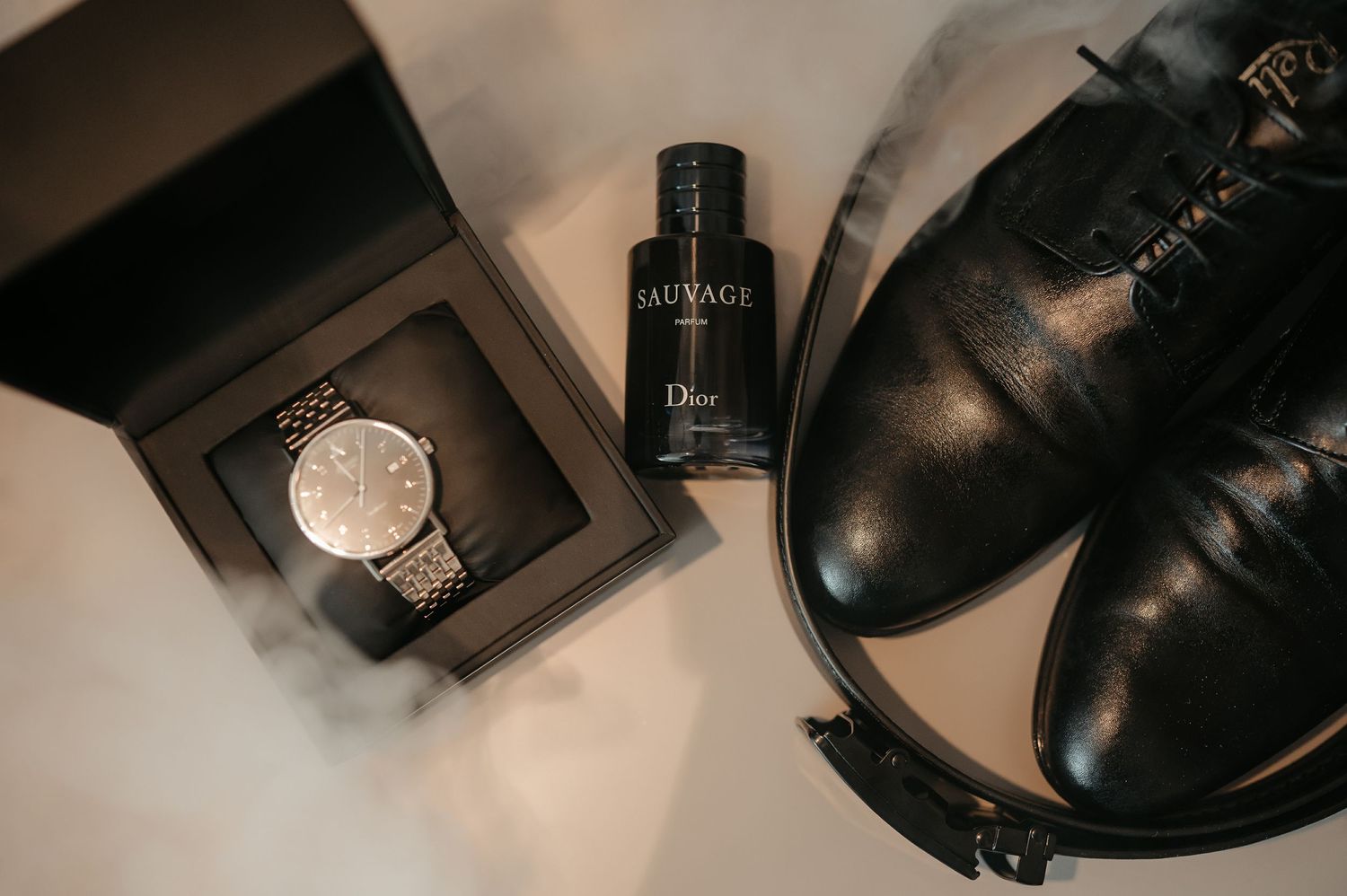 Flat lay of groom accessories including watch, Dior Sauvage cologne, and black leather dress shoes.