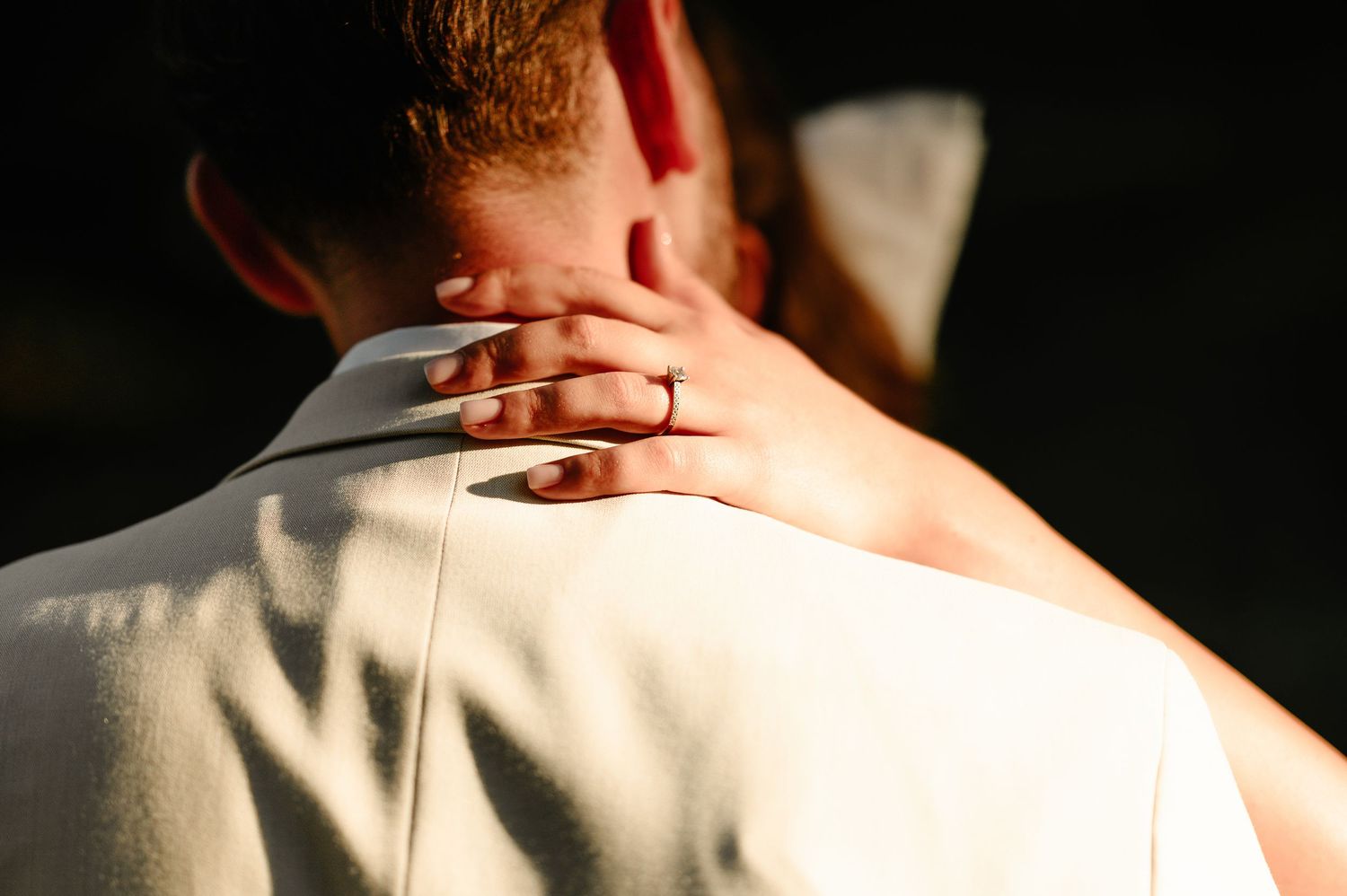 Close-up of brides hand with engagement ring resting on grooms shoulder in warm sunlight.