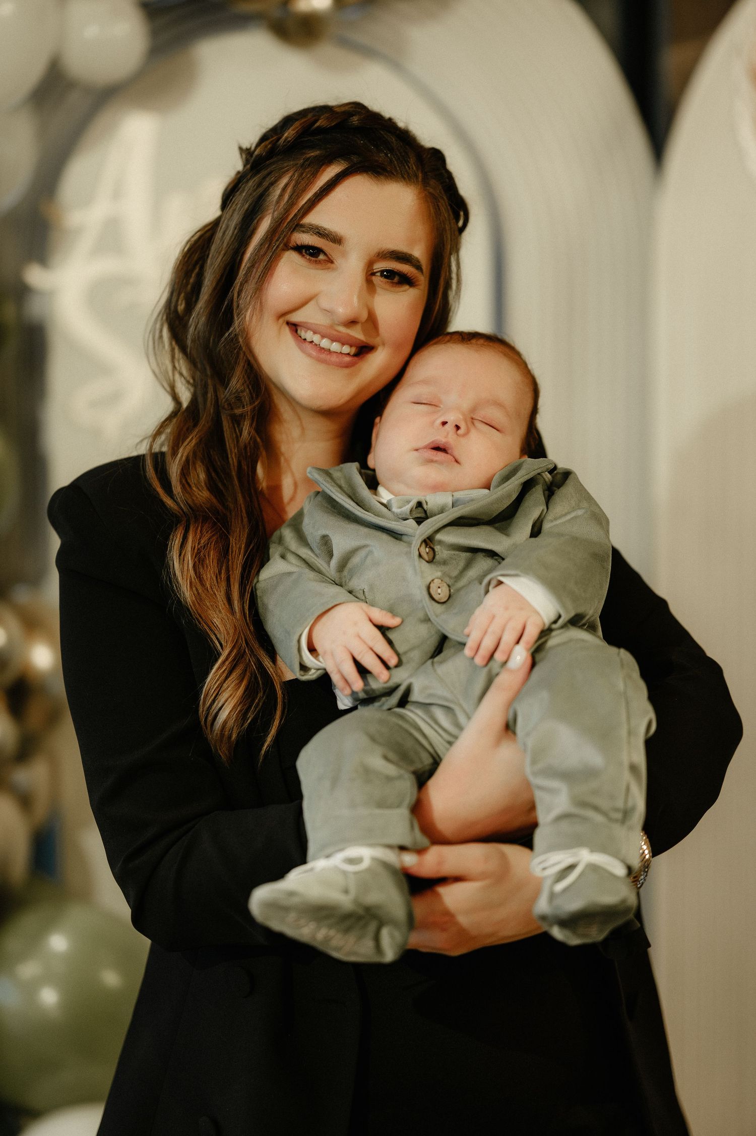 Smiling young mother in black blazer holding sleeping newborn in sage green outfit at baby shower.