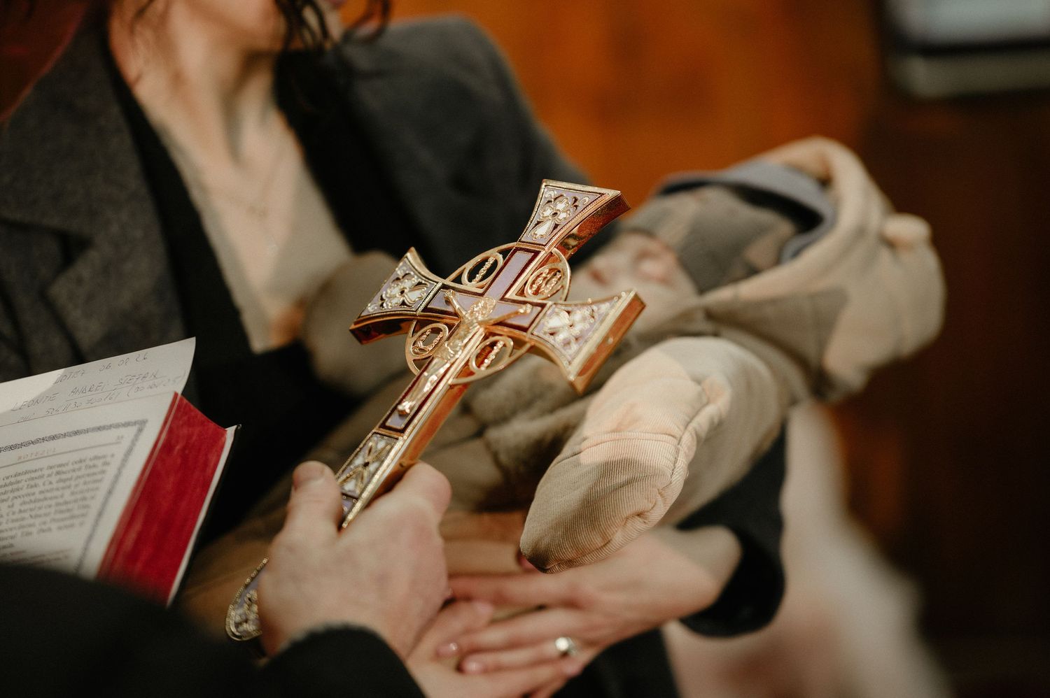 Hands holding an ornate Orthodox Christian cross during a baptism ceremony with a book nearby.