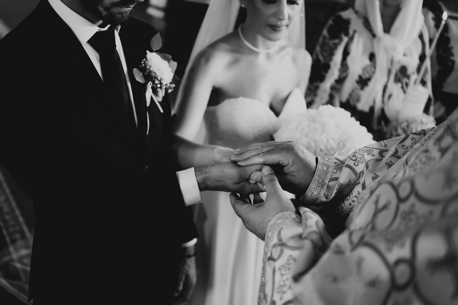 Black and white close-up of Orthodox priest placing wedding rings on couple's hands during ceremony.