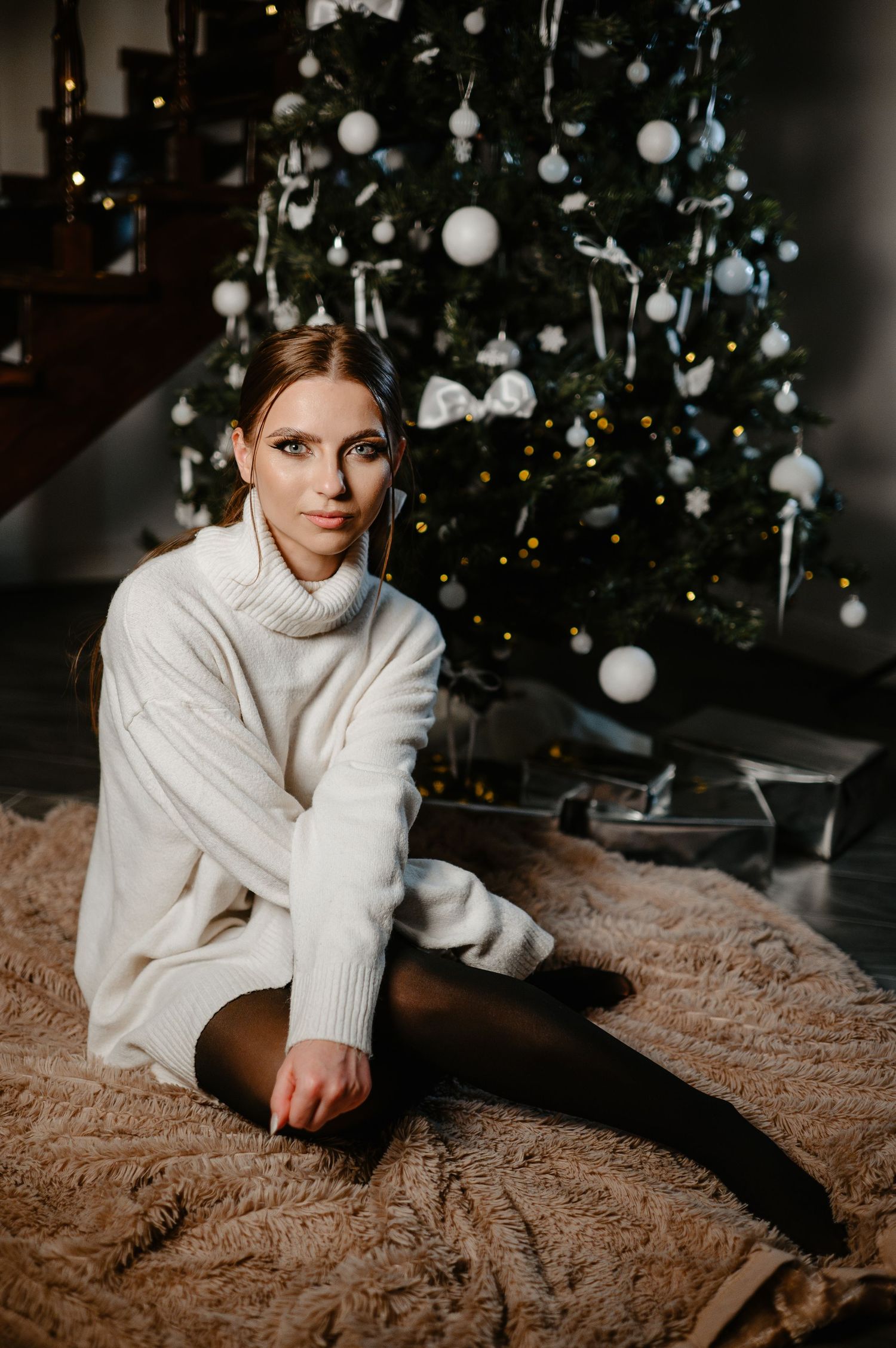 Elegant woman in white turtleneck sweater posing by Christmas tree on fur rug.