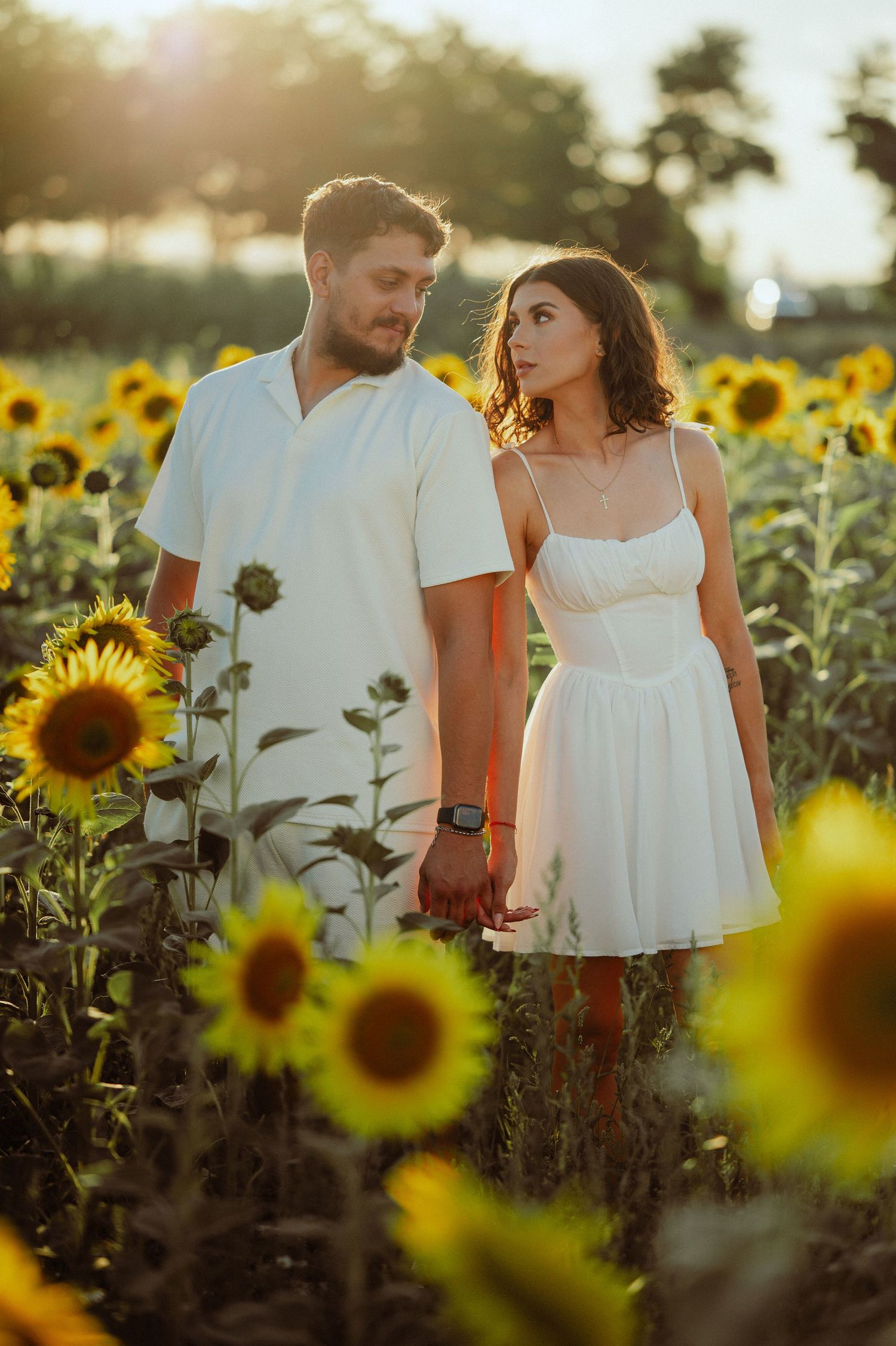 Couple holding hands and walking through a blooming sunflower field in white outfits at sunset.