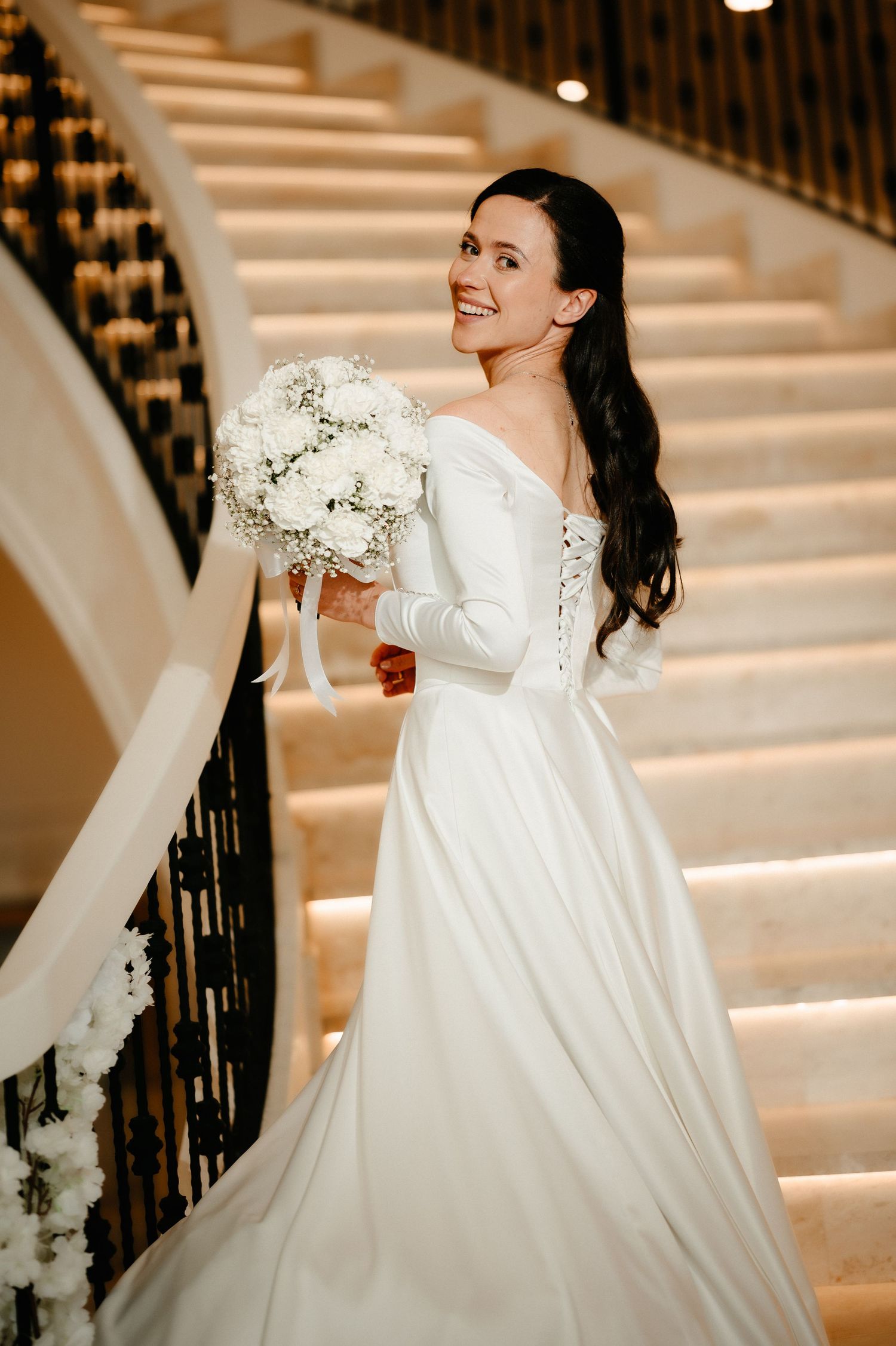 Elegant bride in off-shoulder white gown holding bouquet on grand staircase, smiling.
