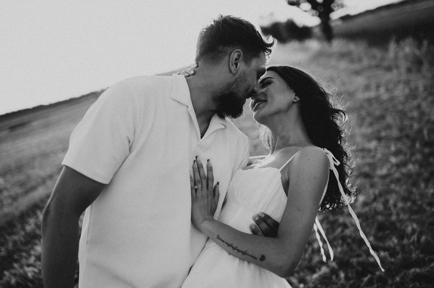 Black and white photo of couple almost kissing in a serene countryside field.