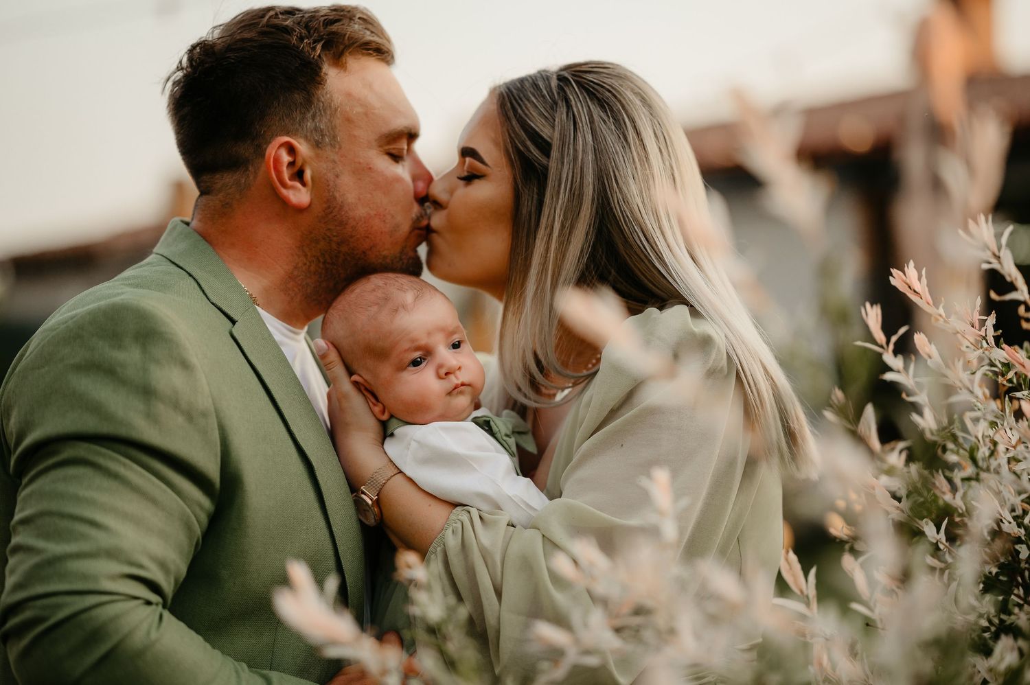 A couple shares a kiss over their newborn baby held between them among soft golden hour foliage.
