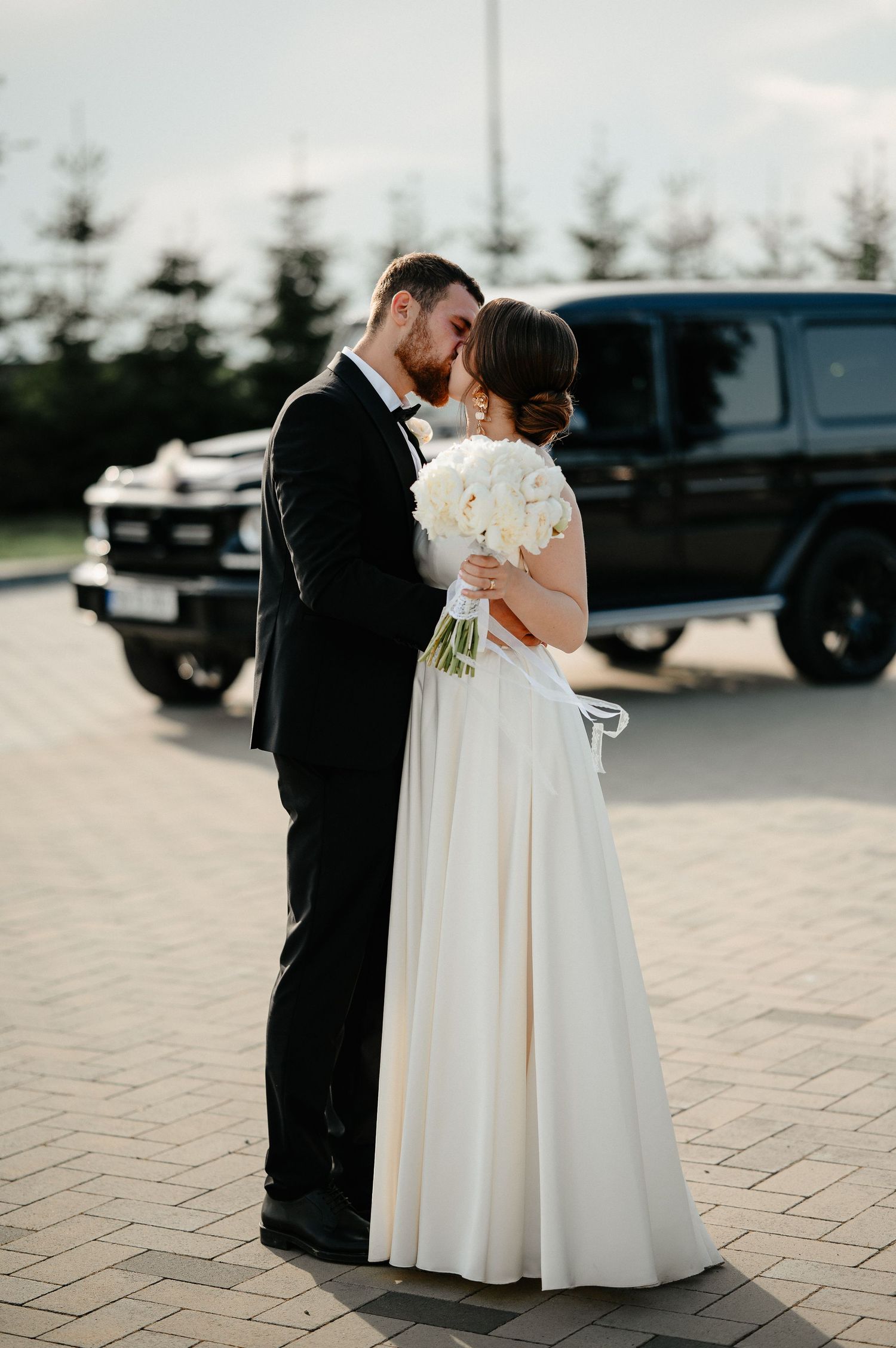 Newlyweds sharing a kiss outdoors with black luxury SUV in background at sunset.