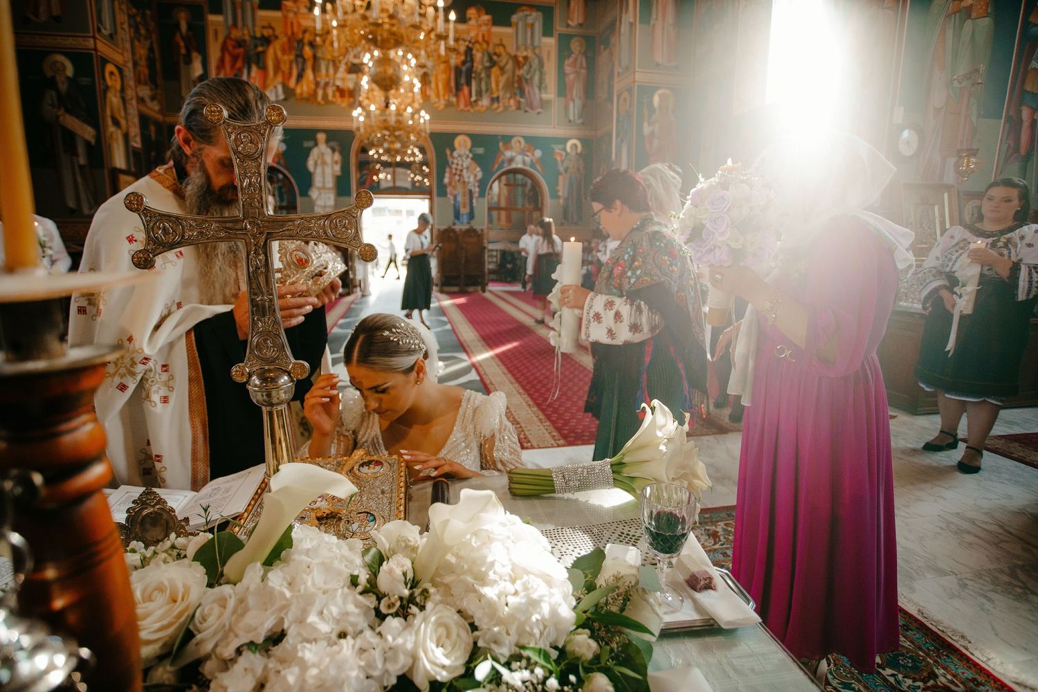 Bride bowing at ornate Orthodox church altar surrounded by white roses and golden candlesticks in sunlit church.