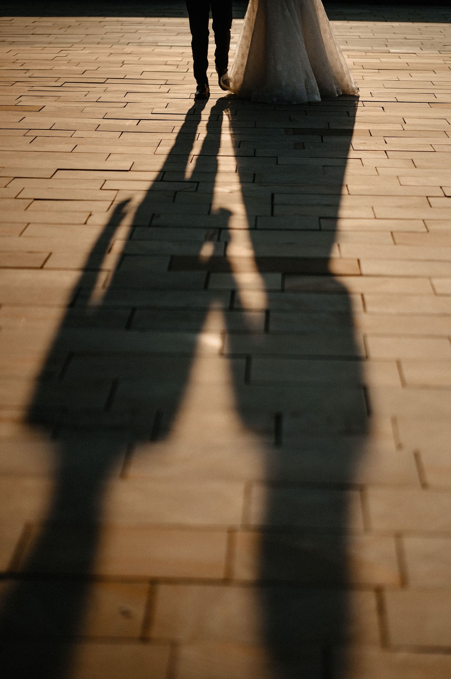 Wedding couple holding hands with dramatic long shadows cast on stone pavement at sunset.