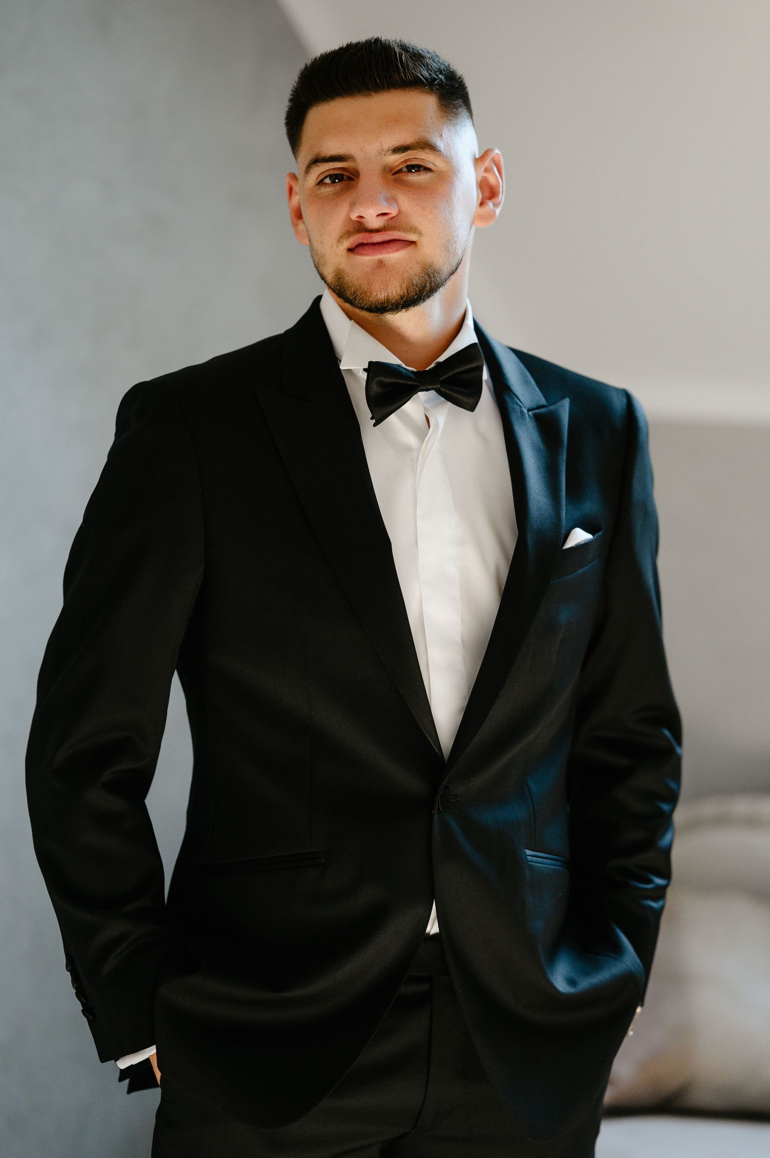 Confident groom in black tuxedo with bow tie and pocket square posing indoors.