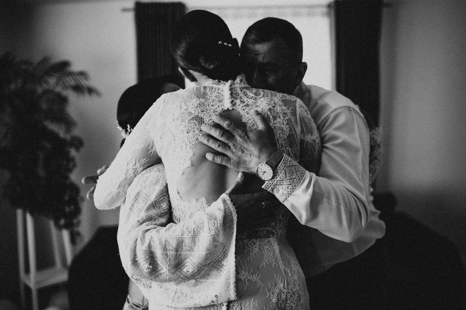 Emotional black and white photo of a bride in lace gown receiving a warm embrace on her wedding day.