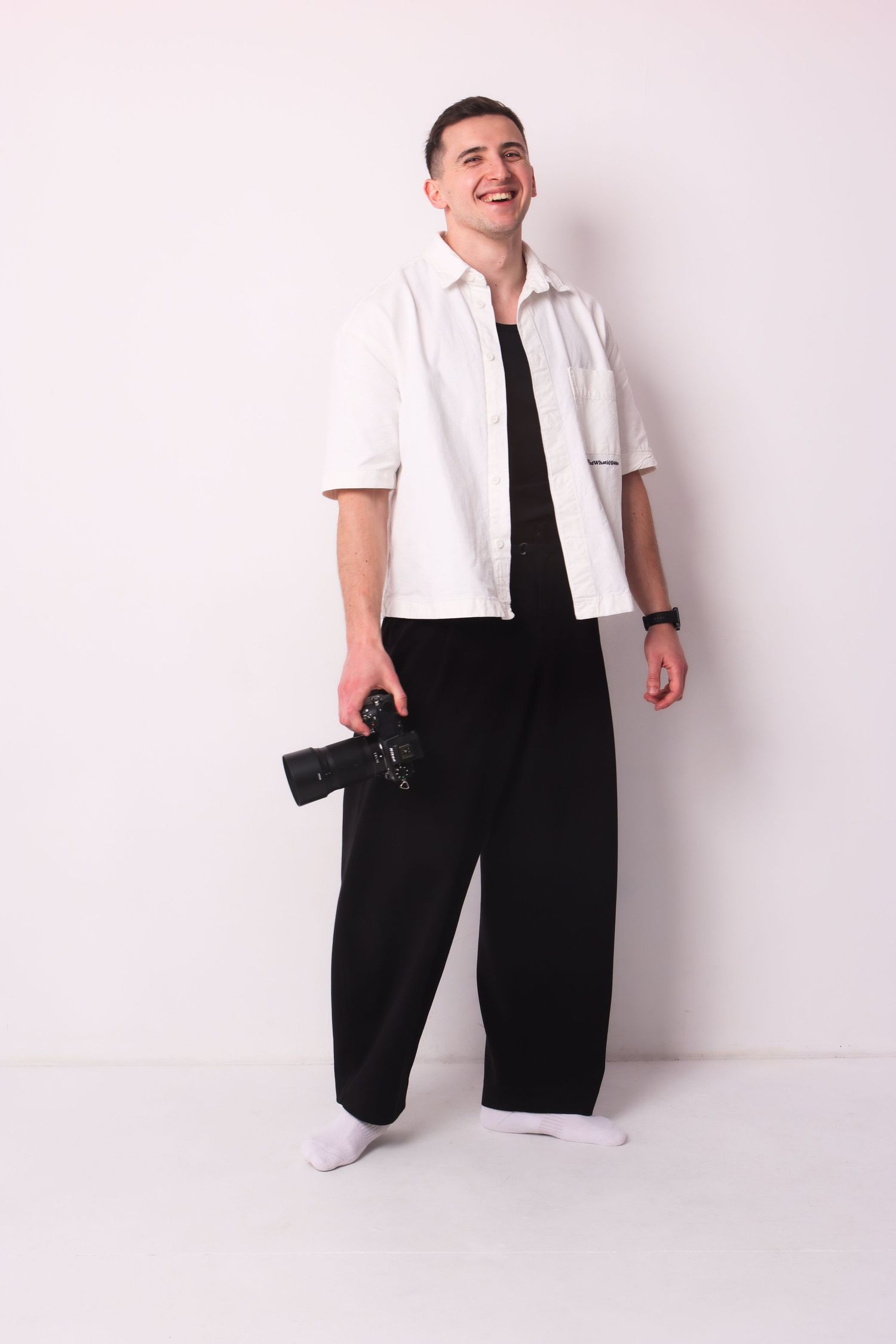 A laughing man in a white shirt holds a camera while posing against a white background.