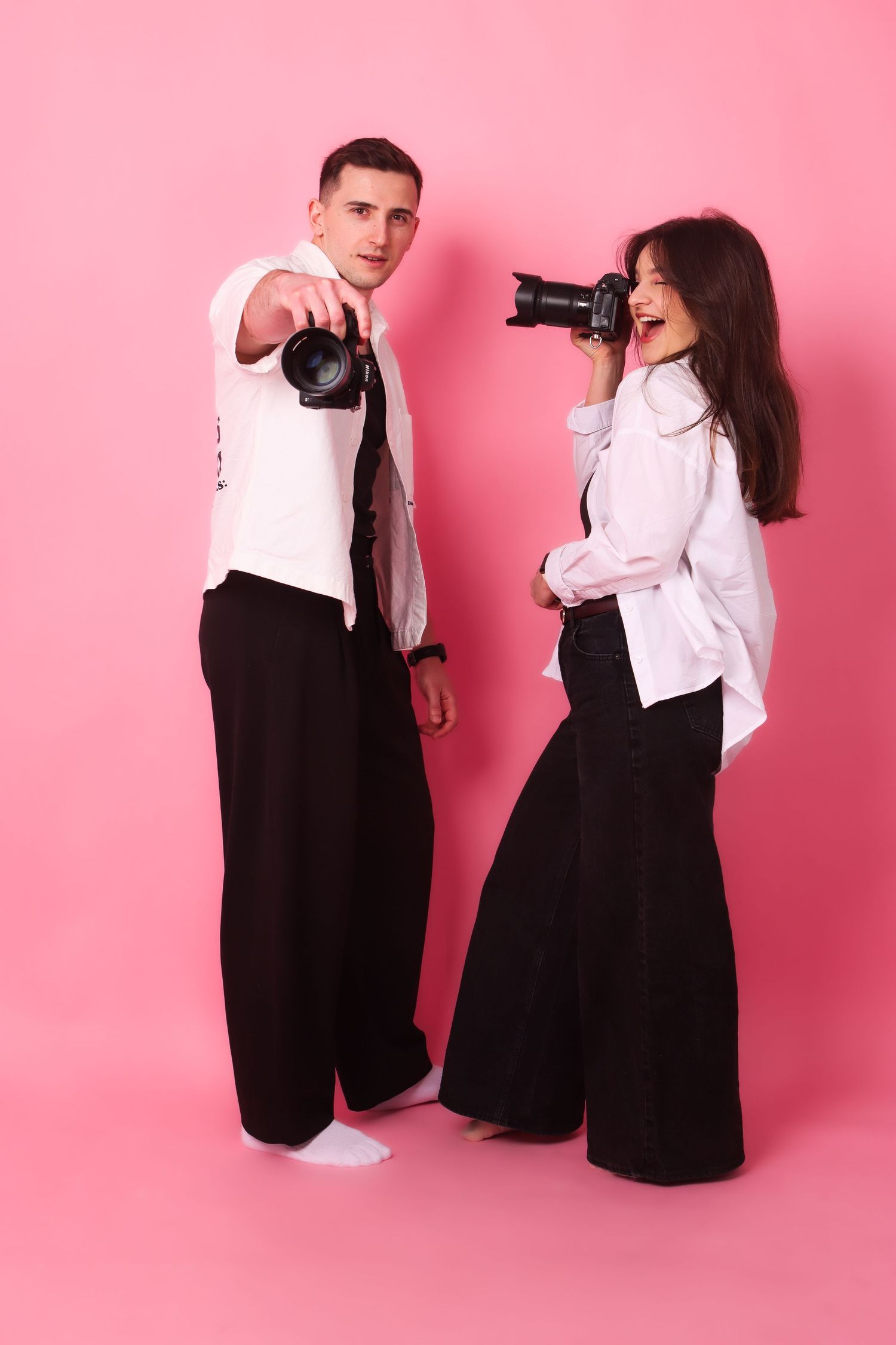 Two photographers in black and white outfits point cameras forward on a pink background.
