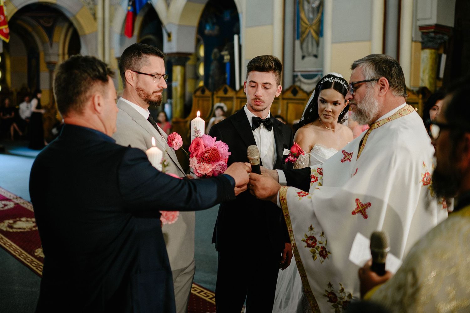 Orthodox priest officiating a wedding ceremony with bride, groom, and groomsmen holding pink flowers.
