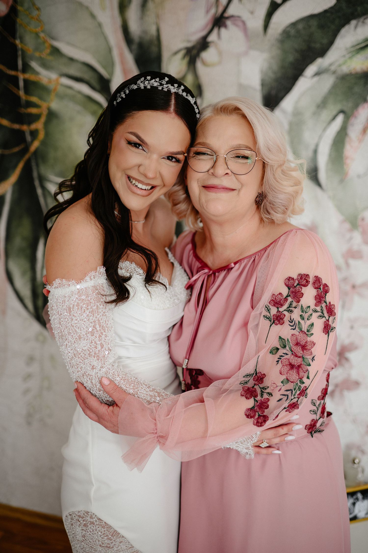 Radiant bride in off-shoulder lace wedding dress smiling and embracing mother in pink floral blouse.