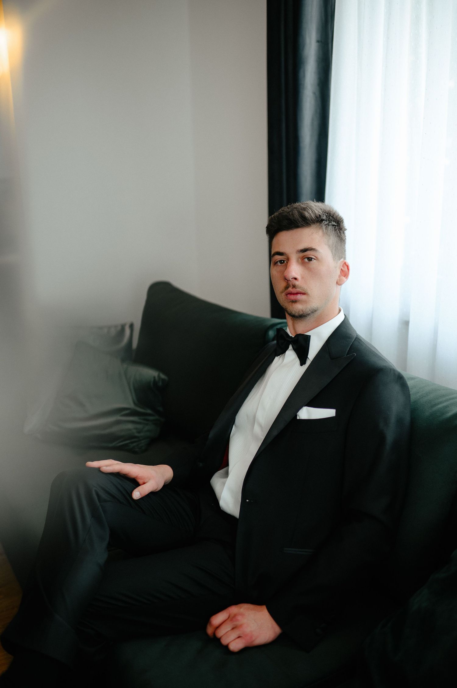 Handsome groom in black tuxedo sitting on green sofa before wedding.