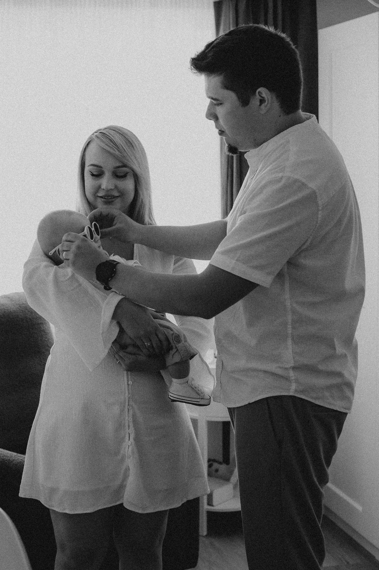 Black and white photo of parents dressing newborn baby together in bright room.
