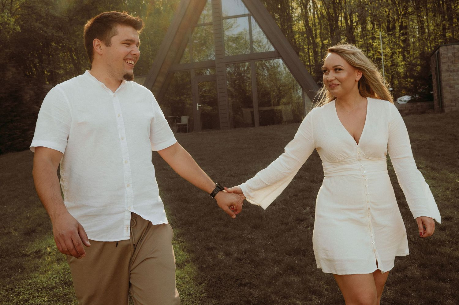 Smiling couple in white outfits holding hands near A-frame cabin in sunlight.