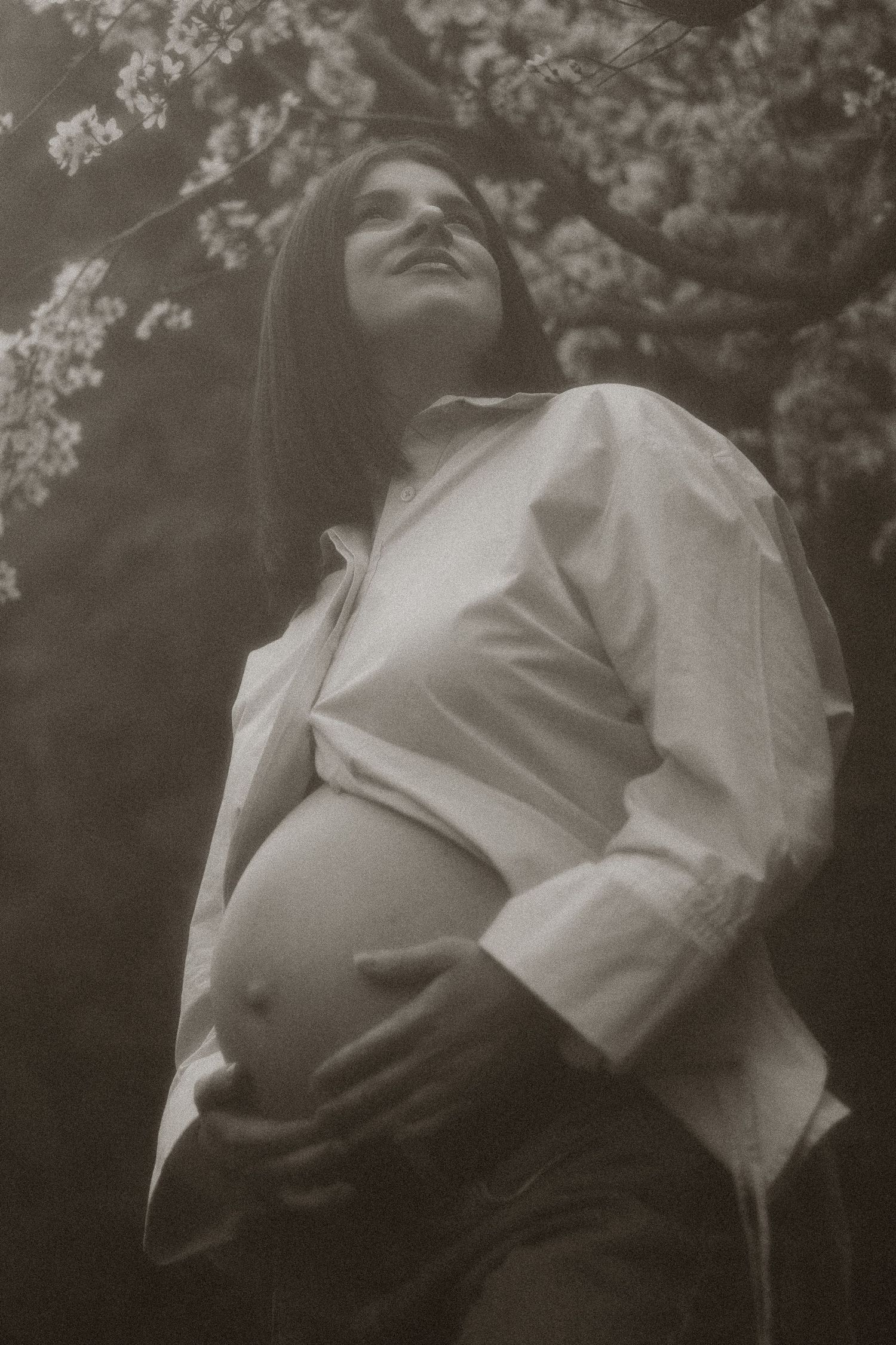 Black and white photo of pregnant woman cradling belly under flowering tree outdoors.