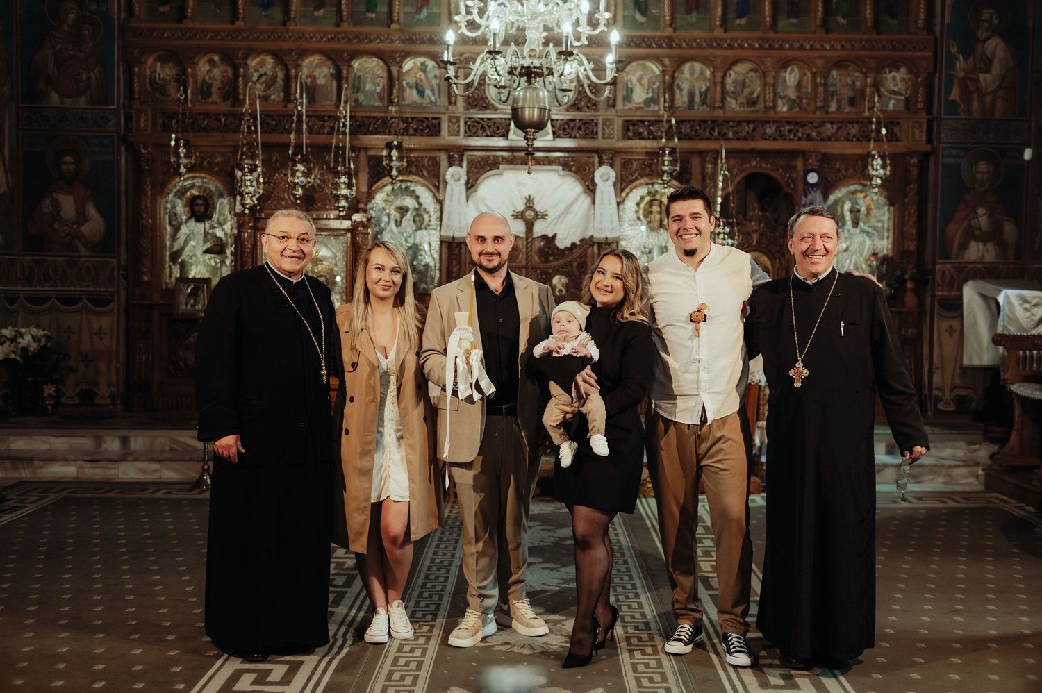 Family group photo with Orthodox priests and baby after a baptism ceremony in a decorated church.