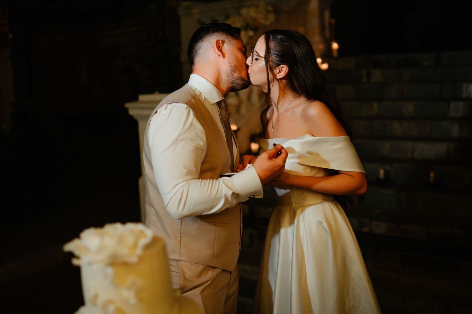 Bride and groom share intimate kiss by candlelight beside white wedding cake in romantic setting.