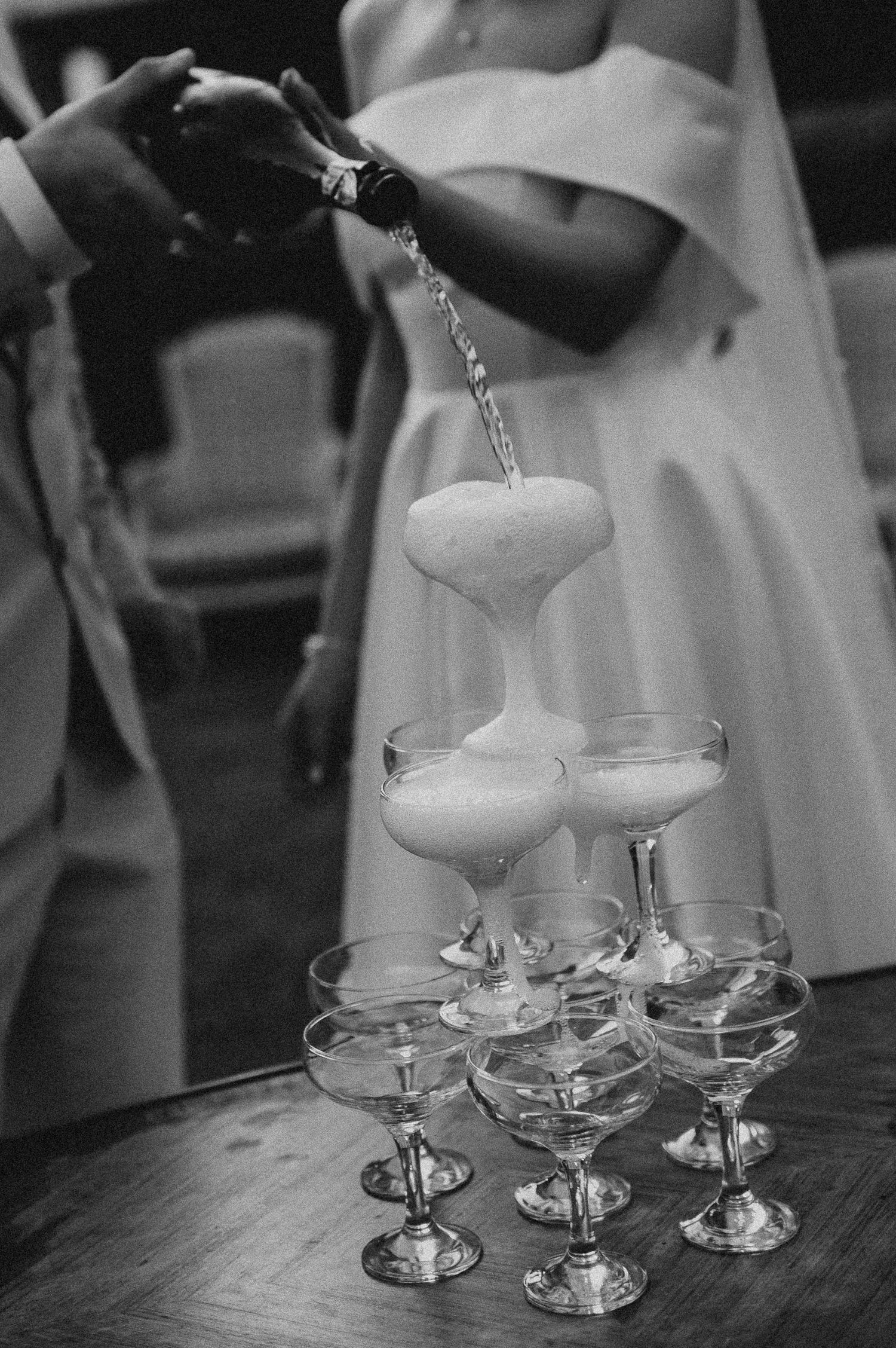 Black and white close-up of champagne being poured into a classic coupe glass tower at a wedding.