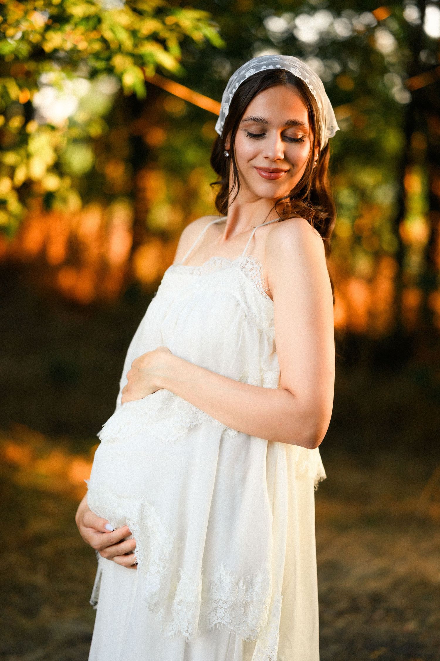 Pregnant woman in white lace dress and veil headpiece cradling baby bump outdoors in golden sunset light.