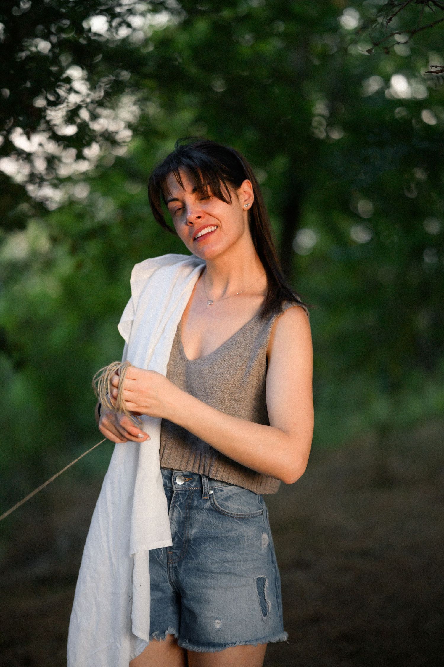 Smiling brunette woman in gray tank top and denim shorts holding white cloth outdoors in green nature setting.