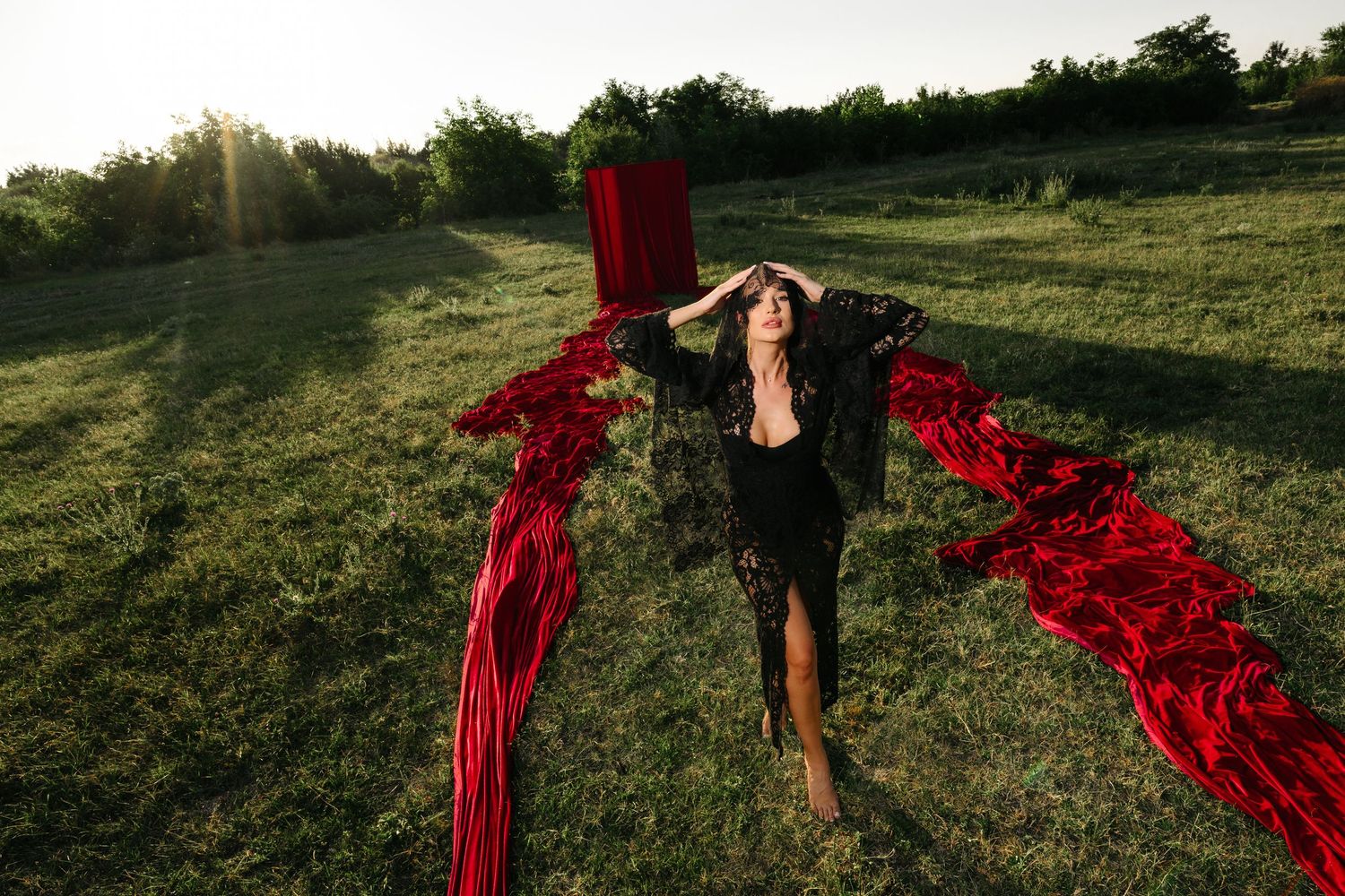 Woman in black dress poses dramatically on green grass with flowing red fabric spread around her in sunlight.