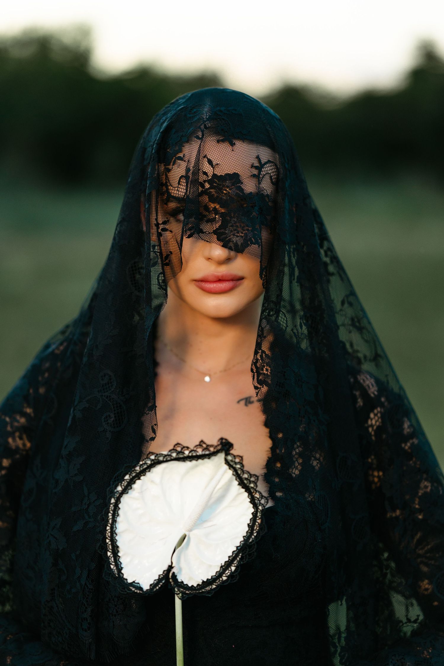 Woman in black lace veil covering eyes holds white anthurium flower outdoors at dusk.